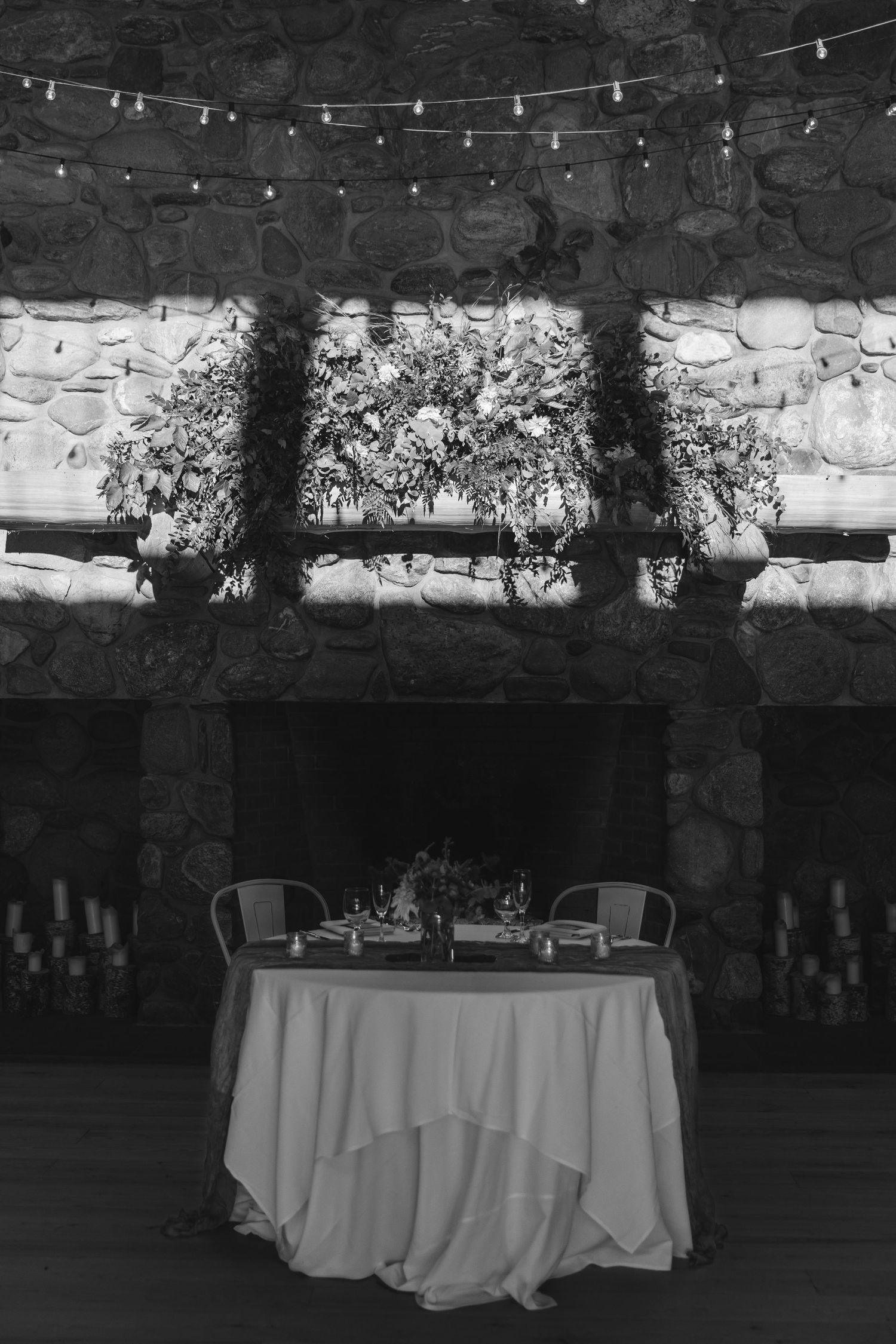 Romantic sweetheart table setup with white tablecloth under string lights and stone wall backdrop.