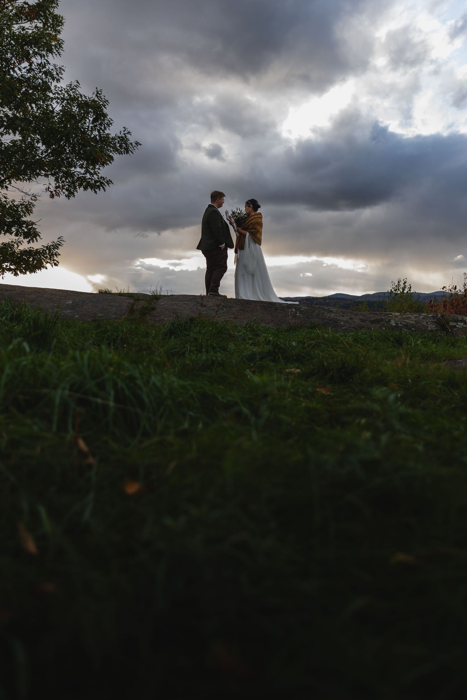 Silhouetted wedding couple standing on hilltop against dramatic cloudy sky.