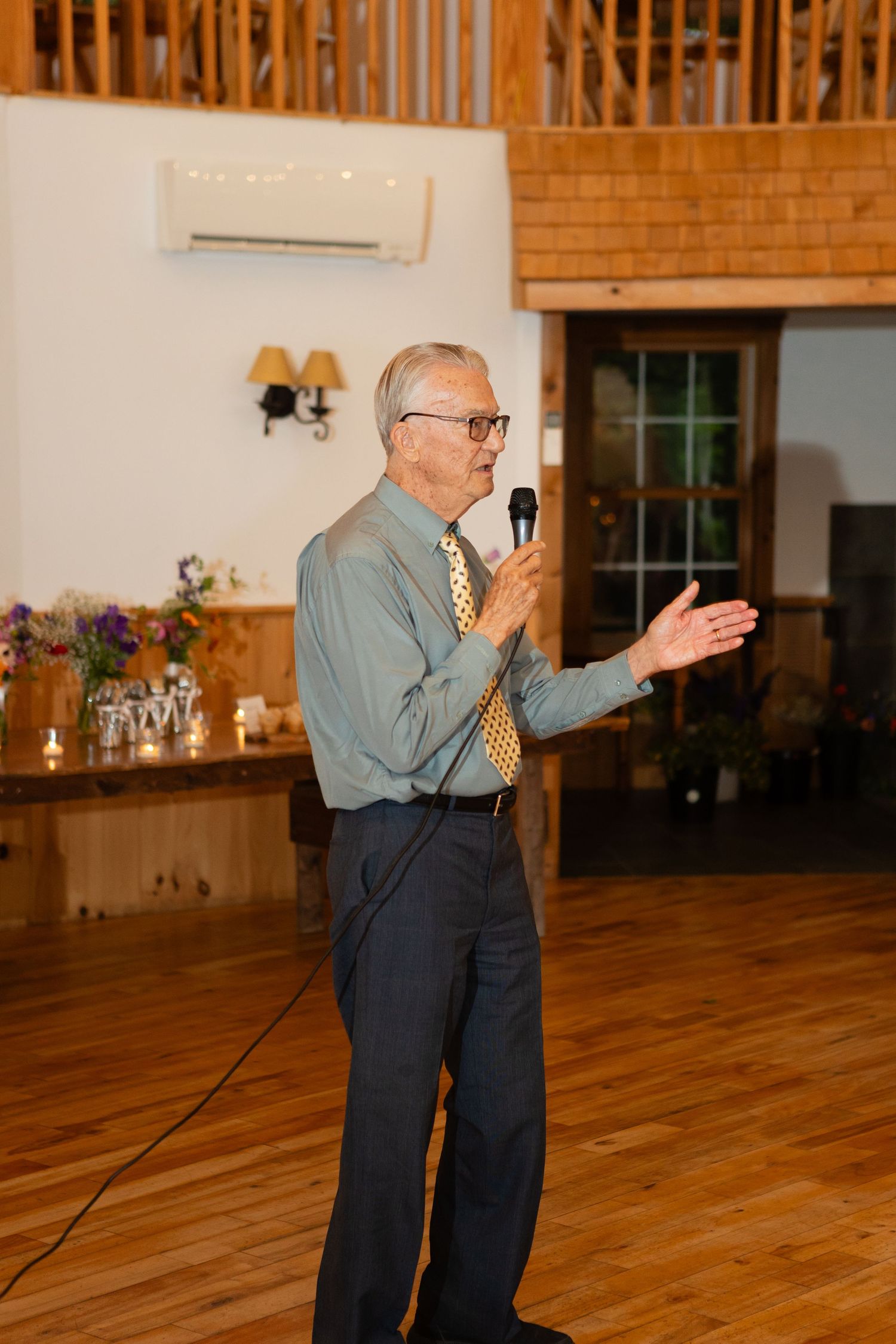 Speaker with microphone addresses guests in rustic wood-paneled reception venue.