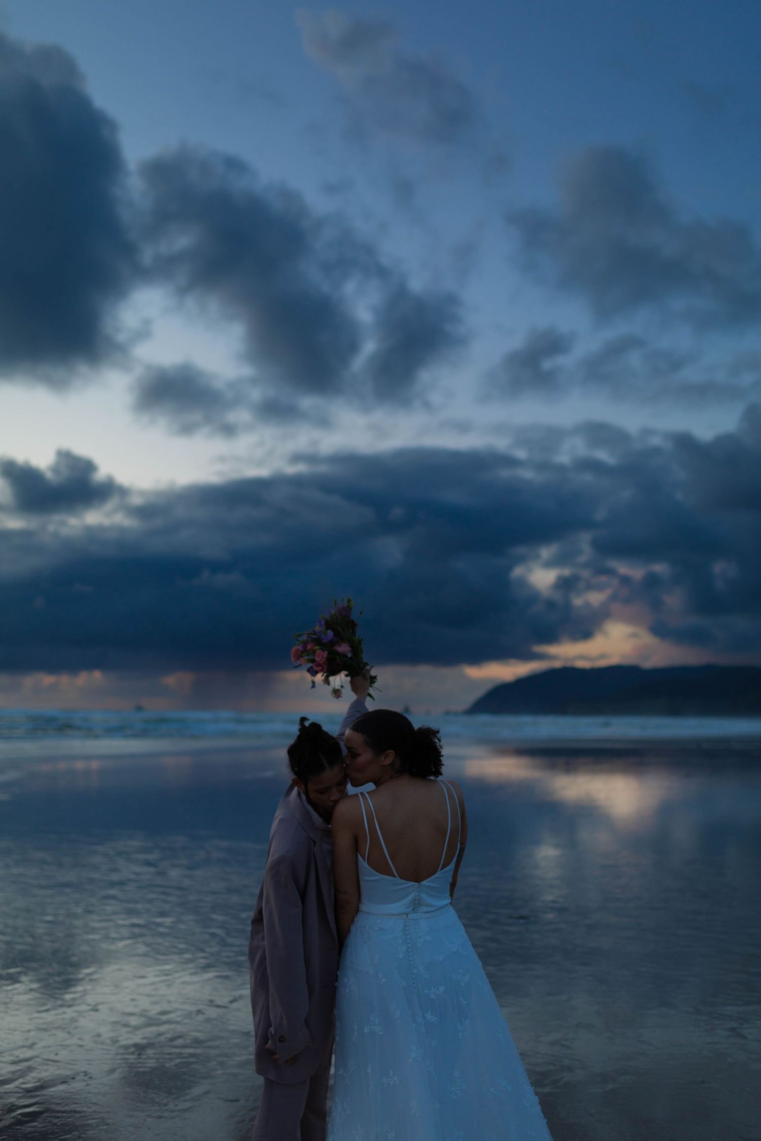 A couple embraces on a beach with dark storm clouds and mountains in the distance.
