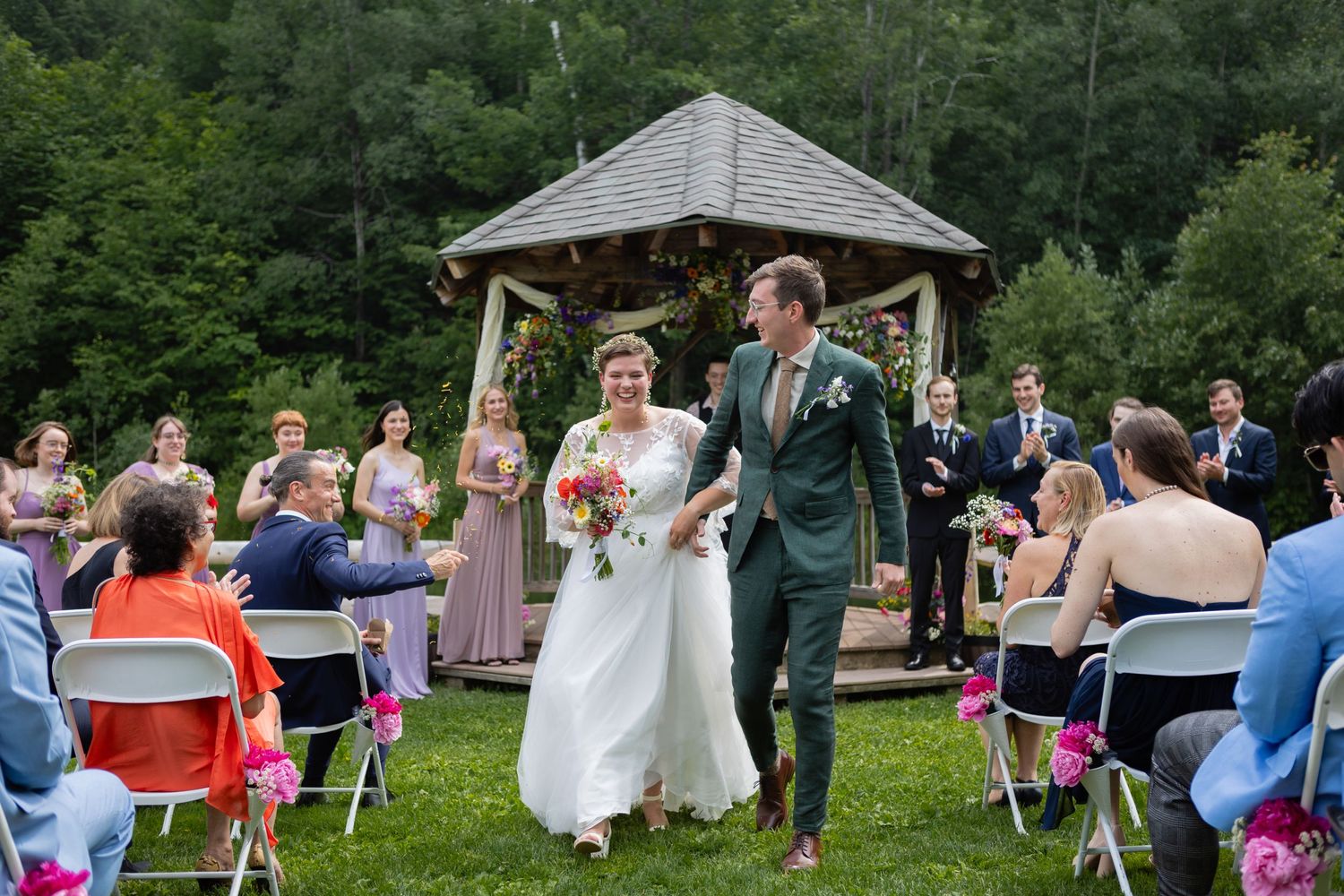 Wedding ceremony takes place at outdoor gazebo with guests seated on folding chairs.