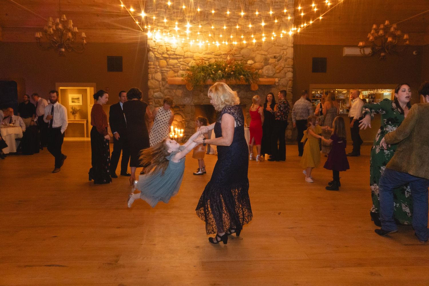 Wedding guests dance and celebrate on a wooden dance floor under warm string lights.