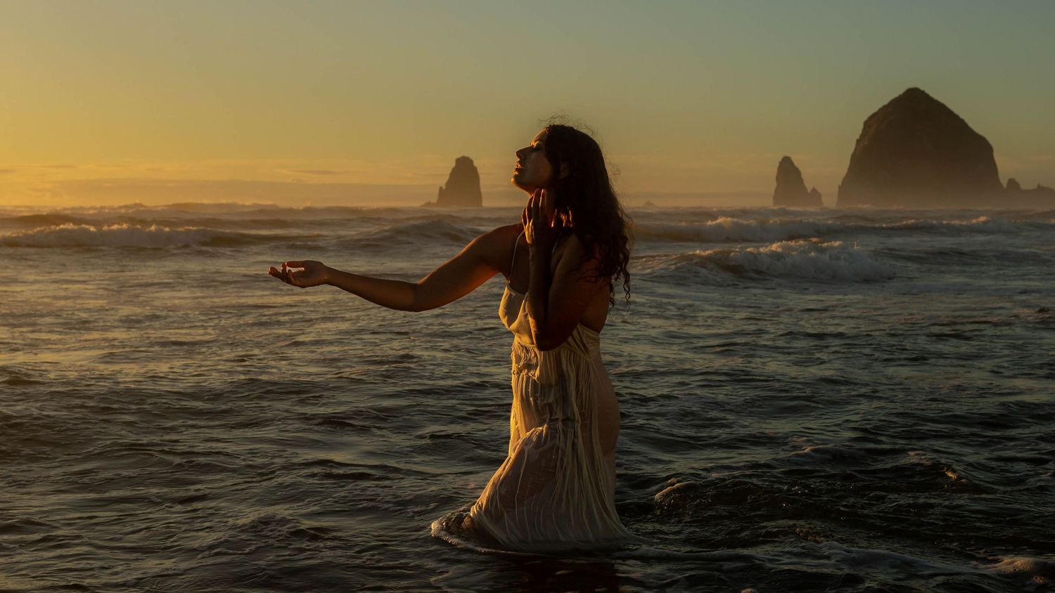 Woman in flowing white dress stands in ocean waves at golden sunset near Haystack Rock.