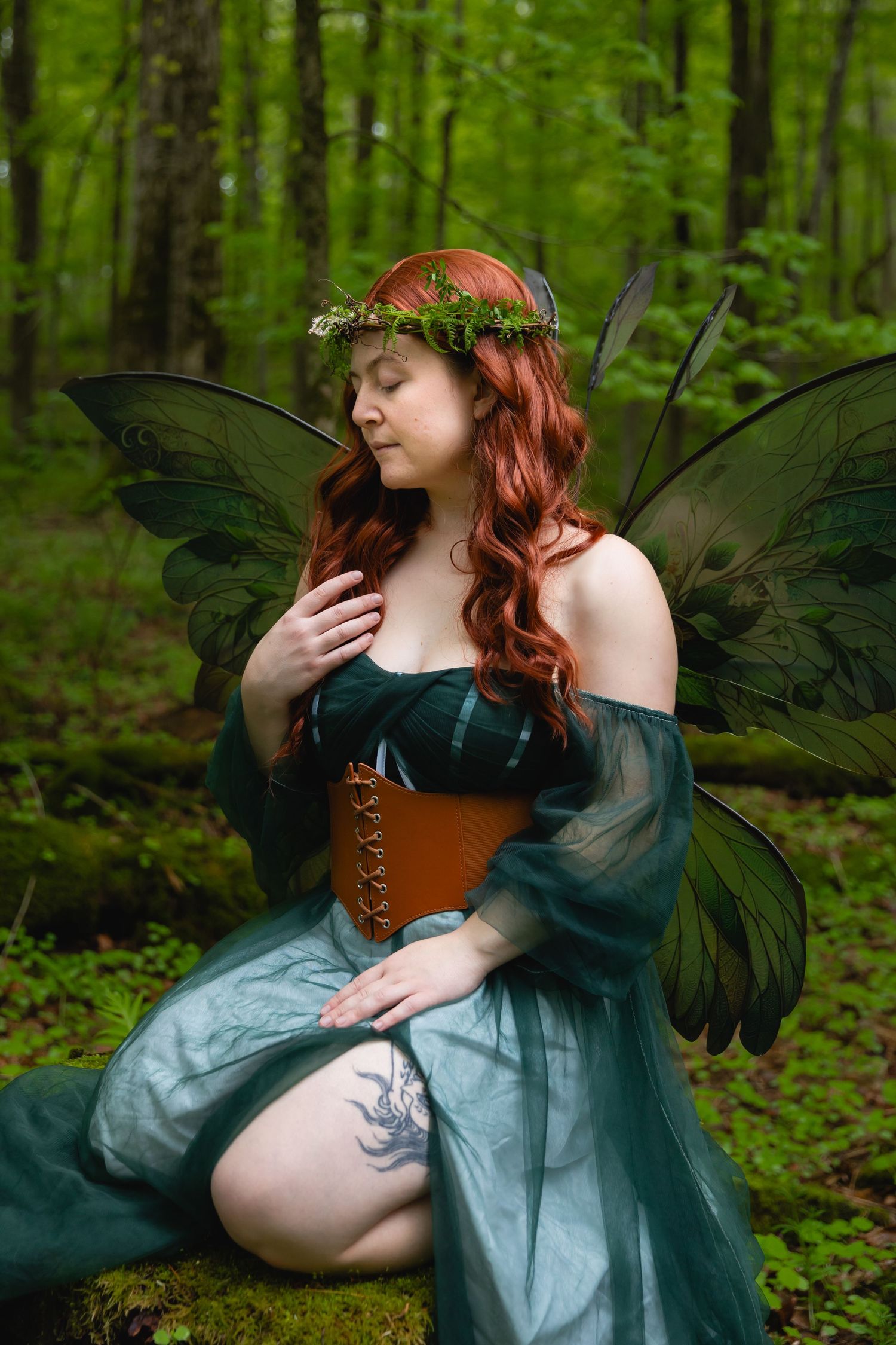 Redheaded woman in teal fairy gown with green wings and floral crown kneels in a lush forest.