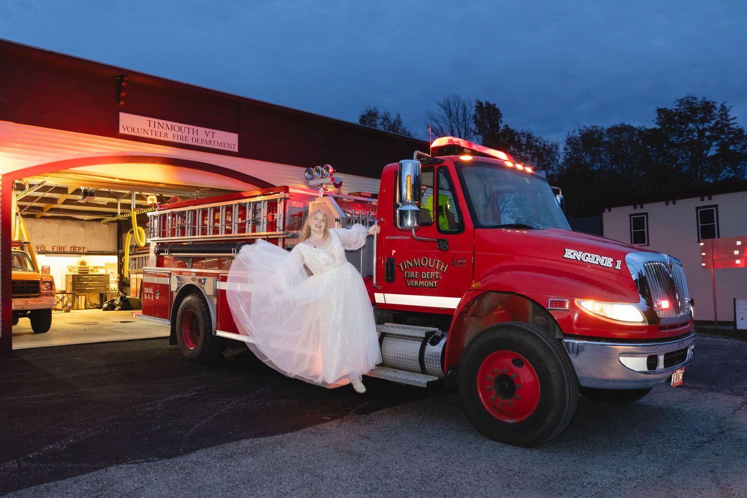 Bride in wedding dress poses next to red fire truck at night outside fire station.