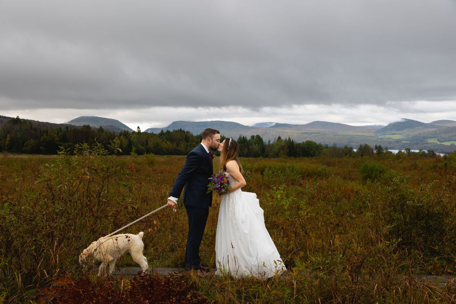 A couple shares a romantic moment with their white dog in a mountain meadow during an overcast wedding day.