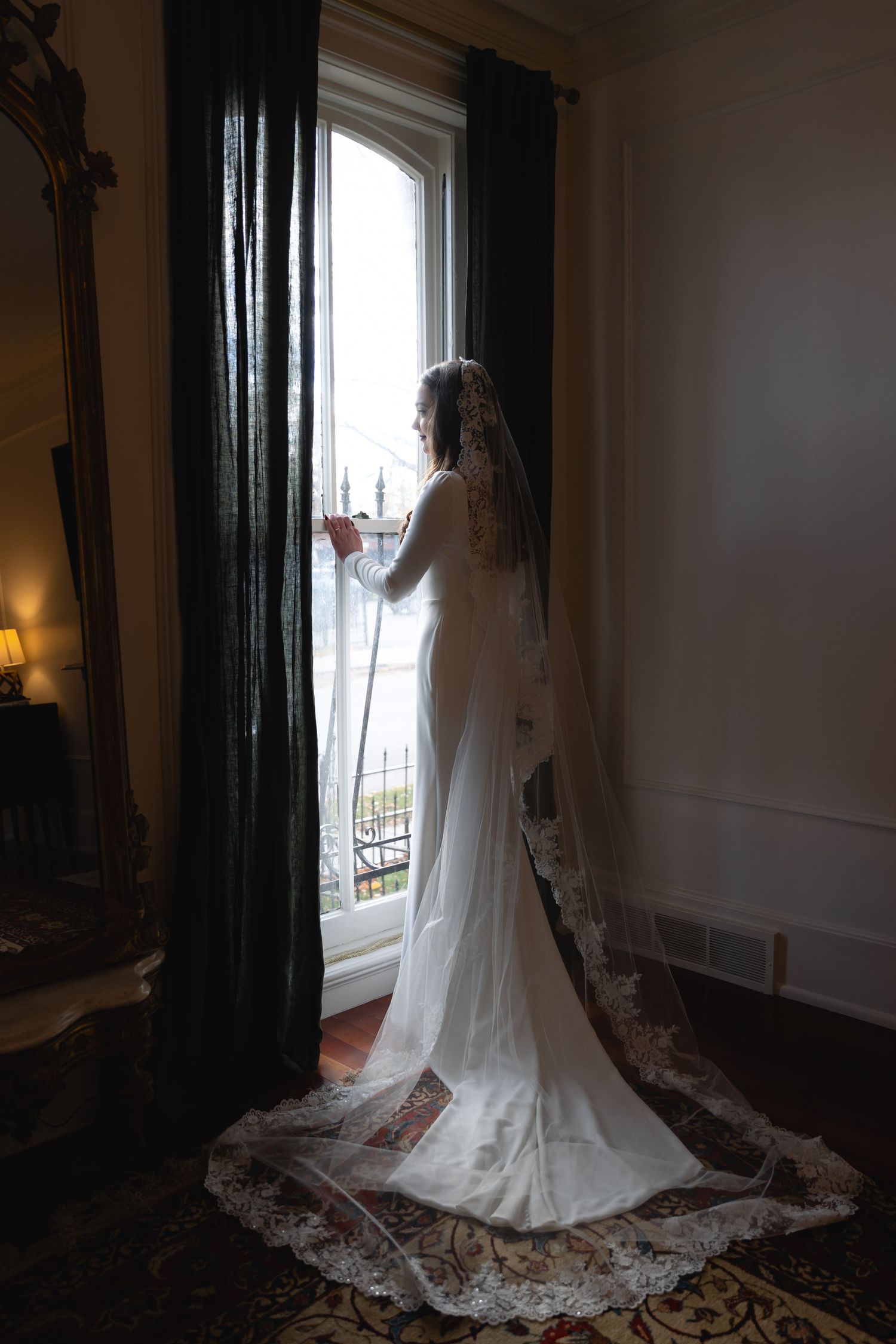 A bride in a flowing white lace gown with long train stands silhouetted against a tall window with dark curtains.