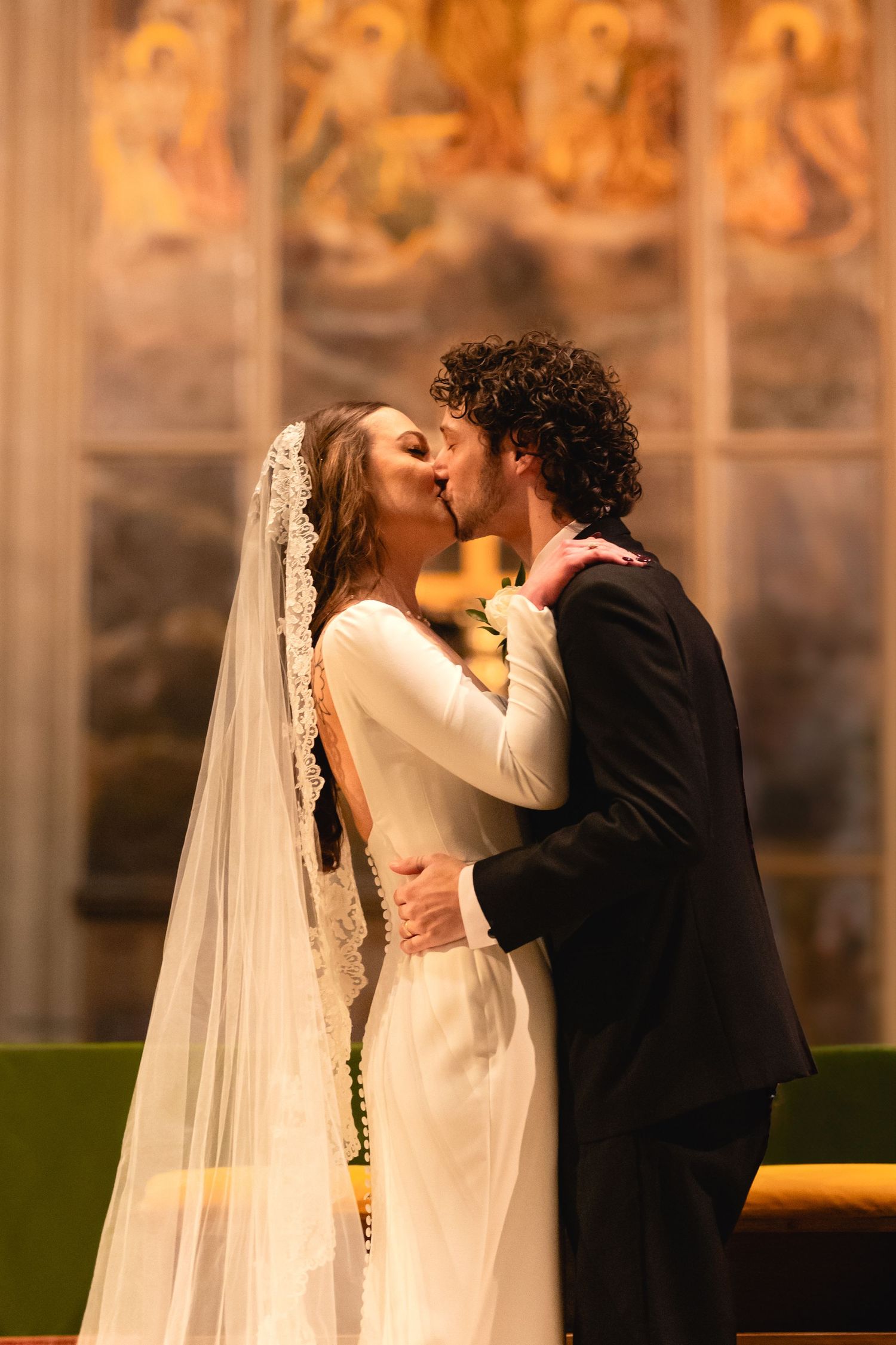A romantic wedding ceremony moment with a couple kissing in a church with ornate decorative panels in the background.