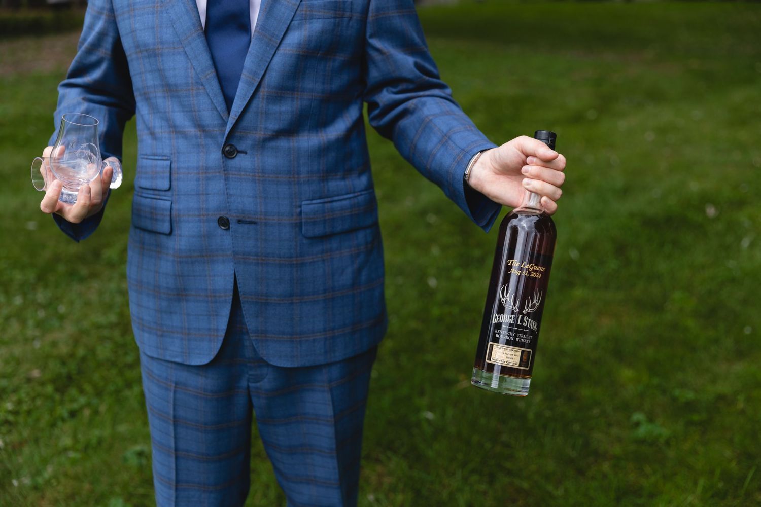 A person in a blue plaid suit holds a bottle of liquor while standing on grass outdoors.