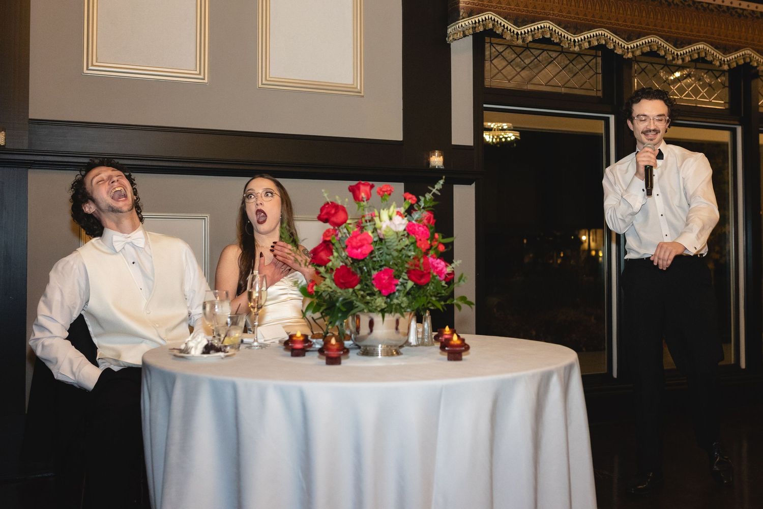 At a wedding reception, people at a sweetheart table share a laugh while listening to a speech.
