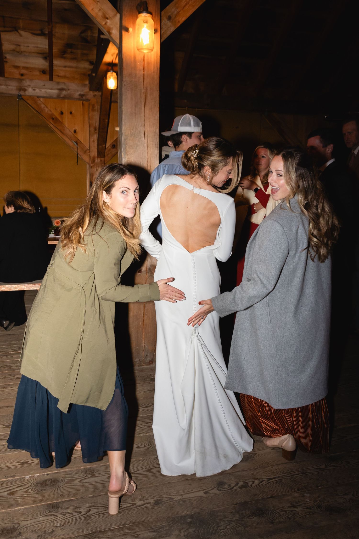 People celebrate at a rustic wedding reception with wood beams and hanging Edison bulbs in a barn setting.