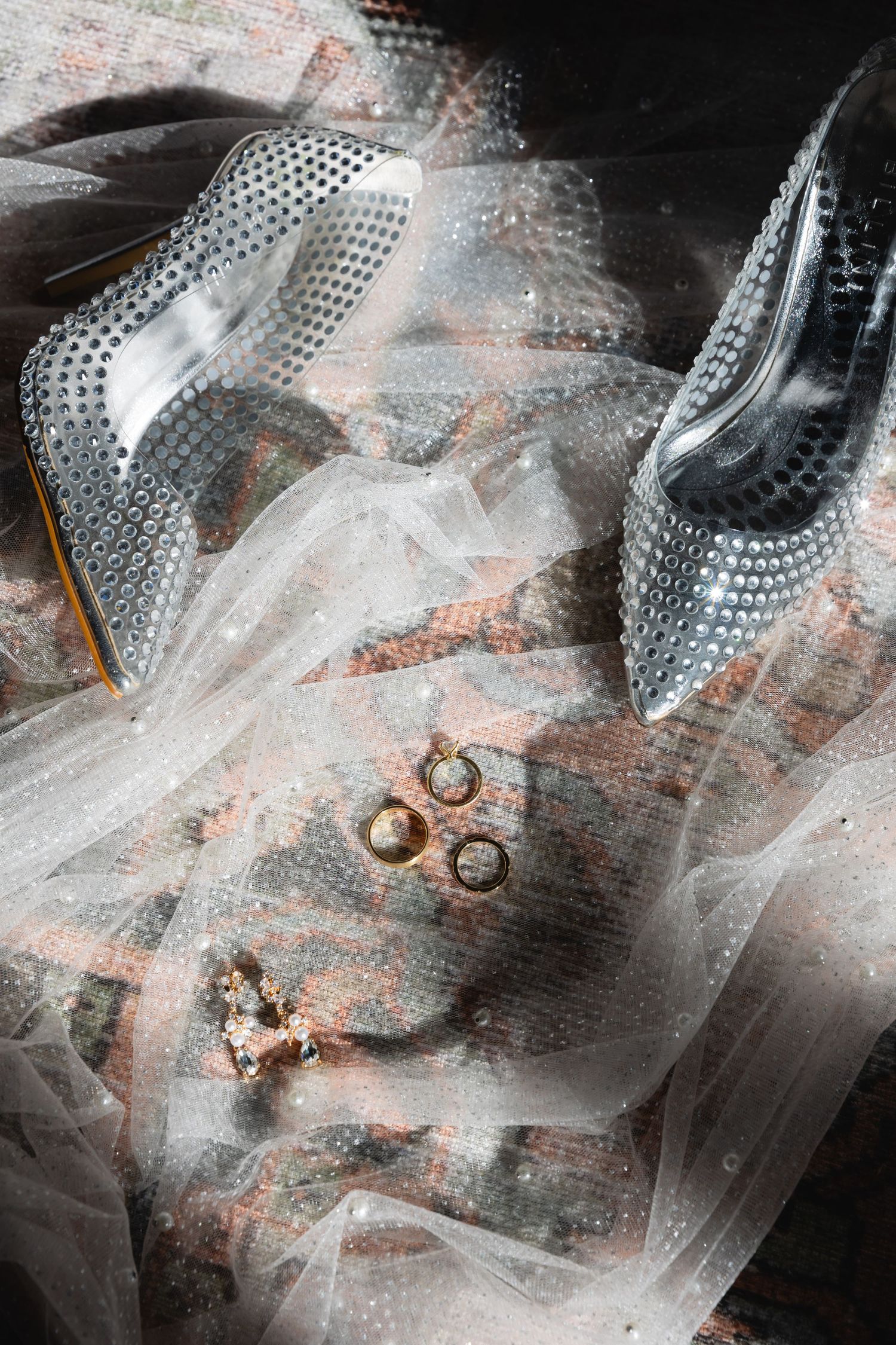 Elegant silver studded bridal shoes with wedding rings and veil draped in soft ethereal light.