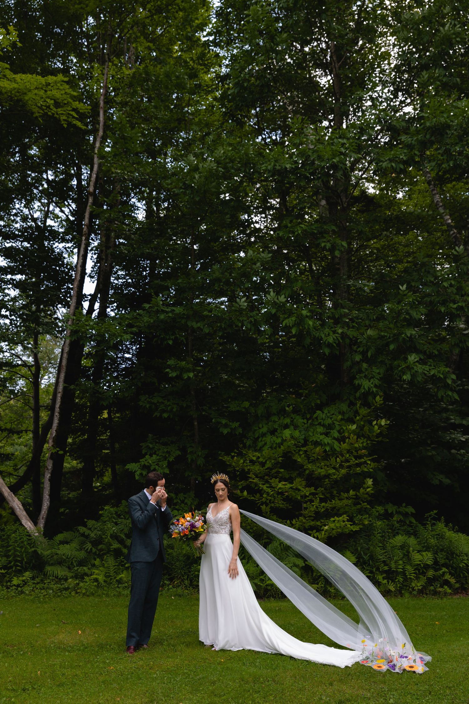 Couple in wedding attire standing in forest clearing with flowing white veil.