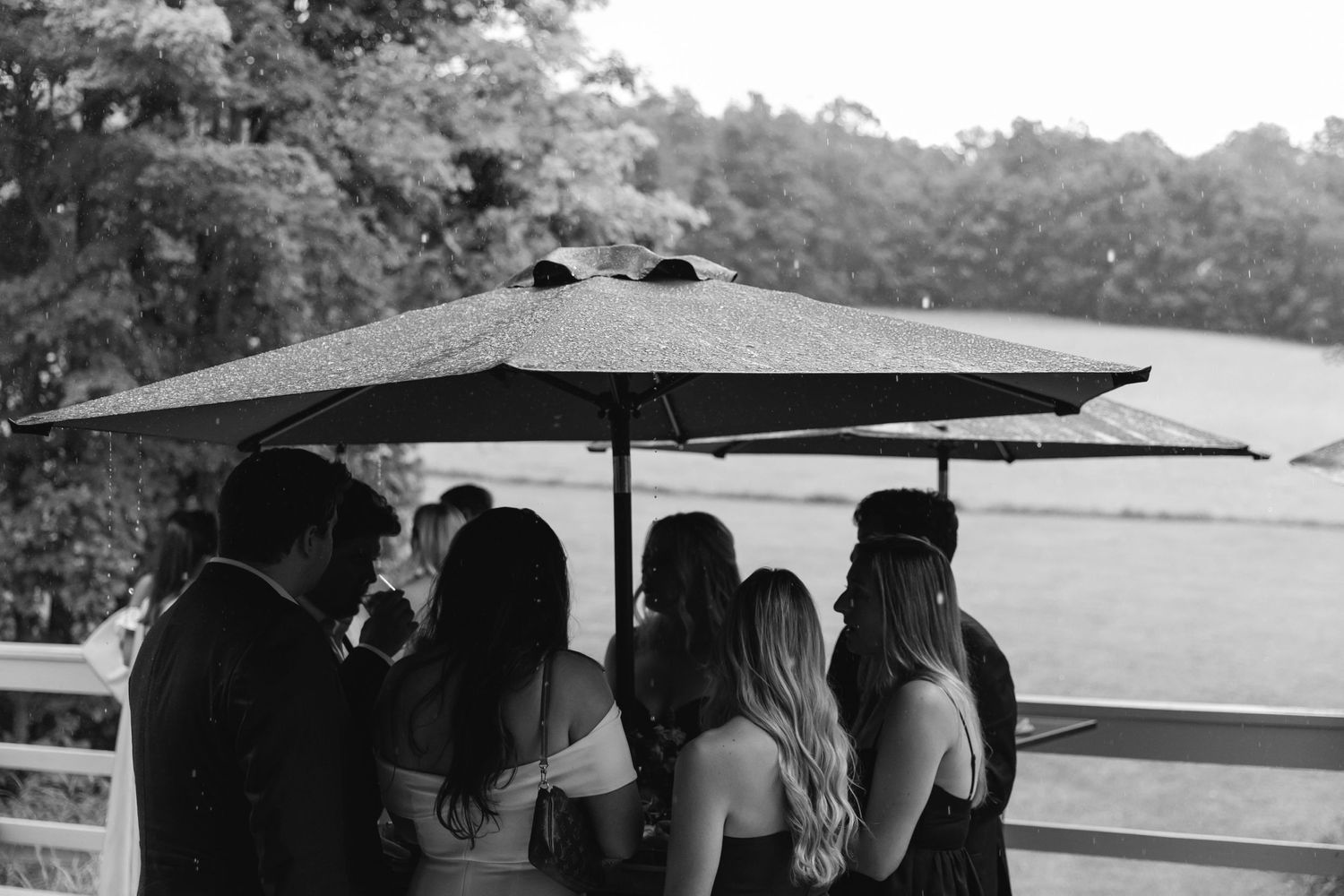 A group of people gather under a large black umbrella on a deck overlooking water in black and white.