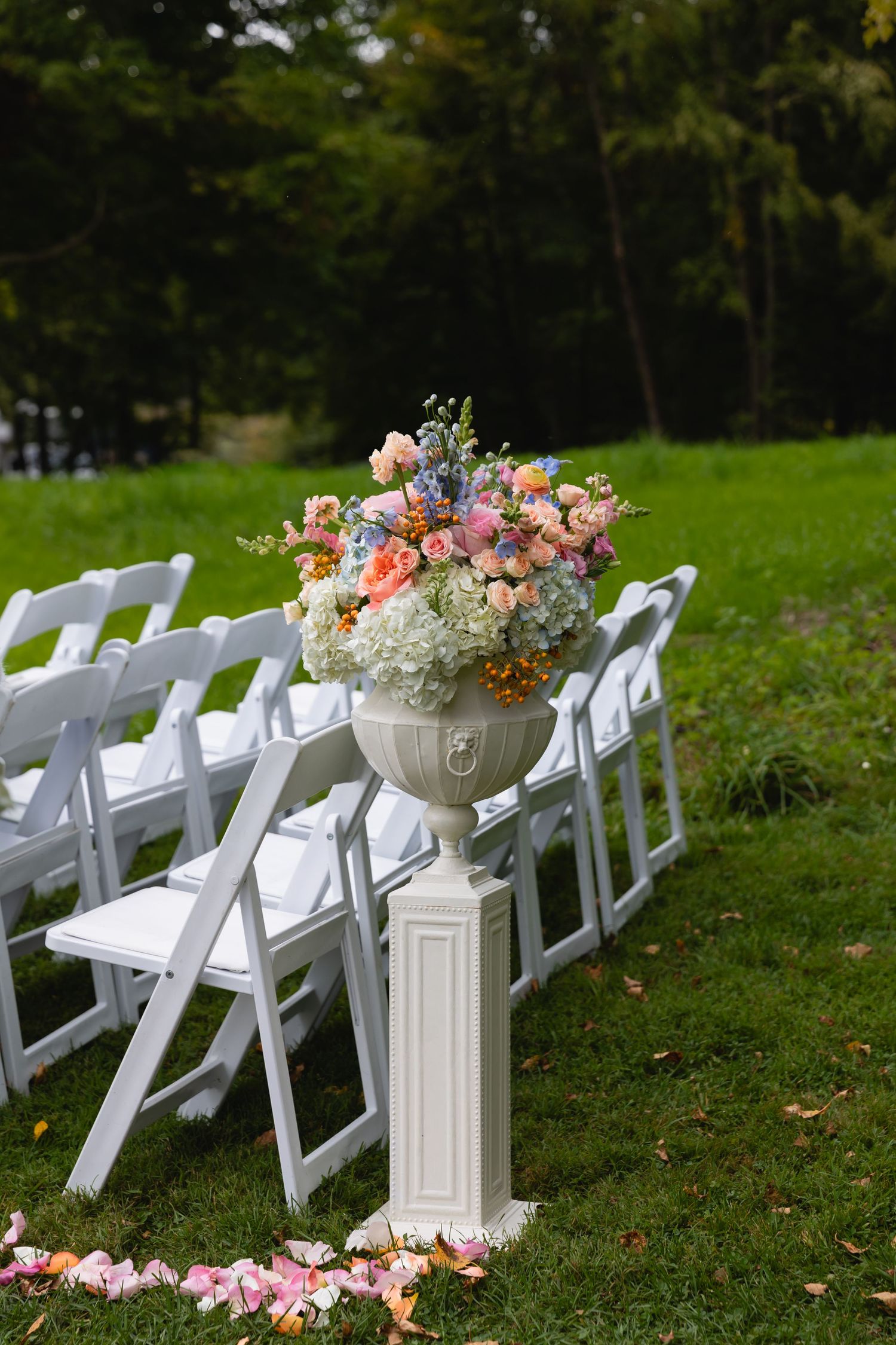 Beautiful floral arrangement in white urn on pedestal next to white folding chairs at outdoor wedding ceremony setup.