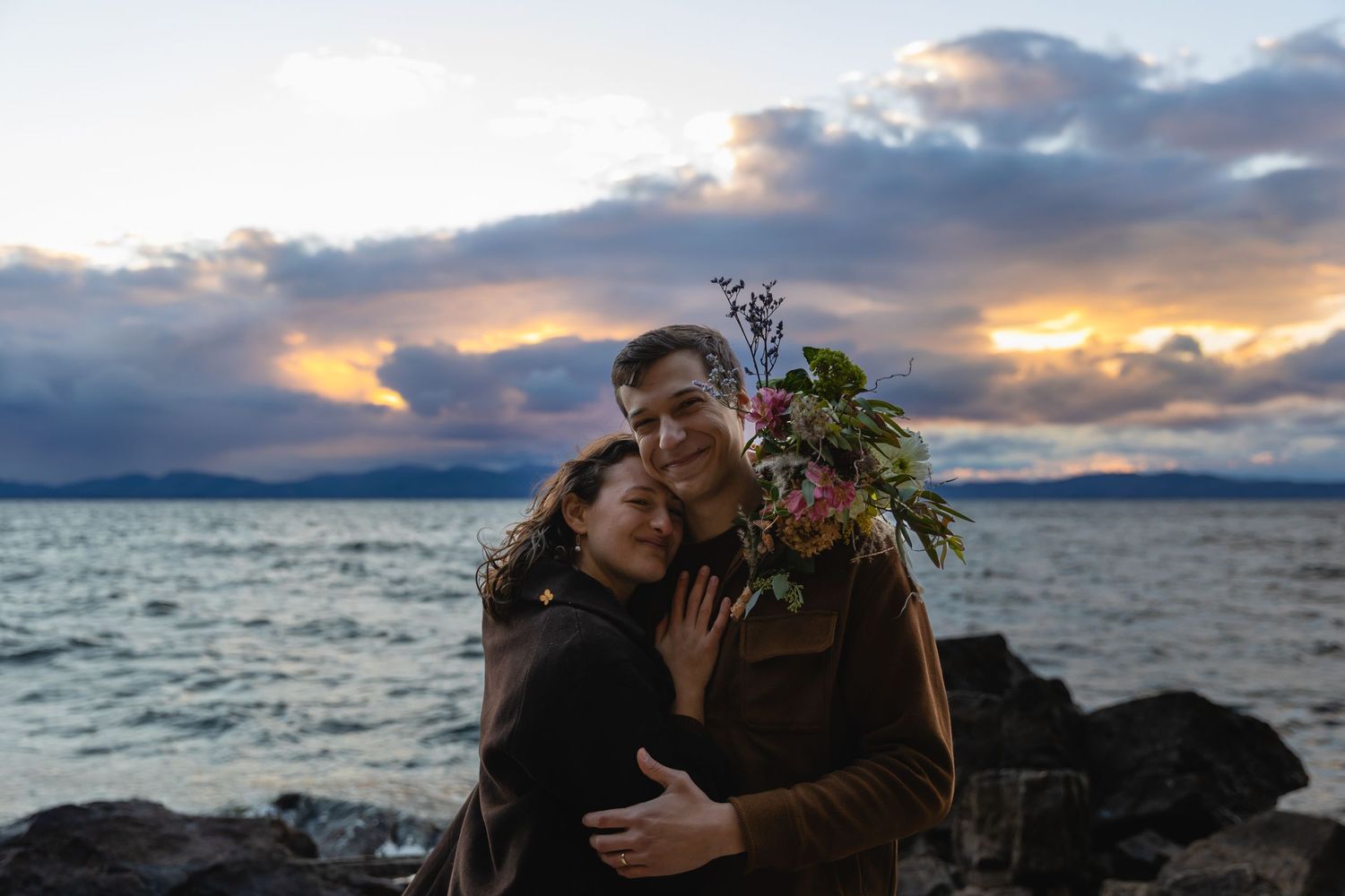 A romantic couple embraces on a rocky beach at sunset with dramatic clouds and ocean waves in the background.