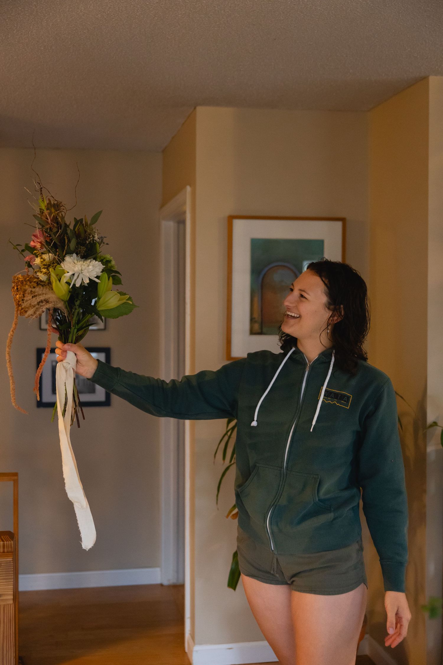 Someone wearing a green hoodie holds up a floral bouquet with white lilies and orange accents in a home interior.