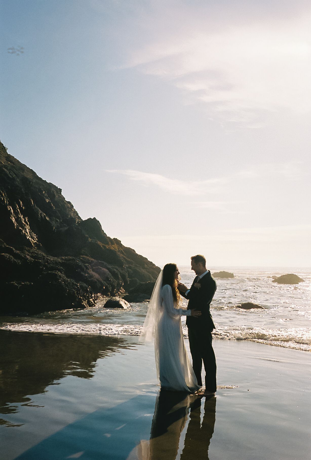 Bride and groom holding hands on wet beach at golden hour with rocky cliffs behind.