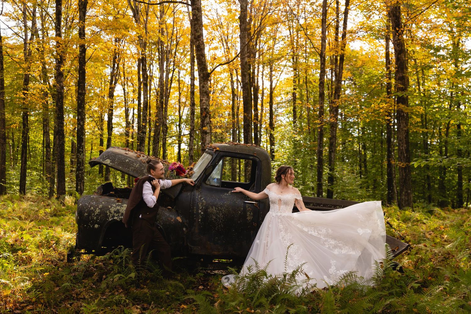 A bride and groom pose beside a rustic old truck surrounded by vibrant yellow fall foliage.