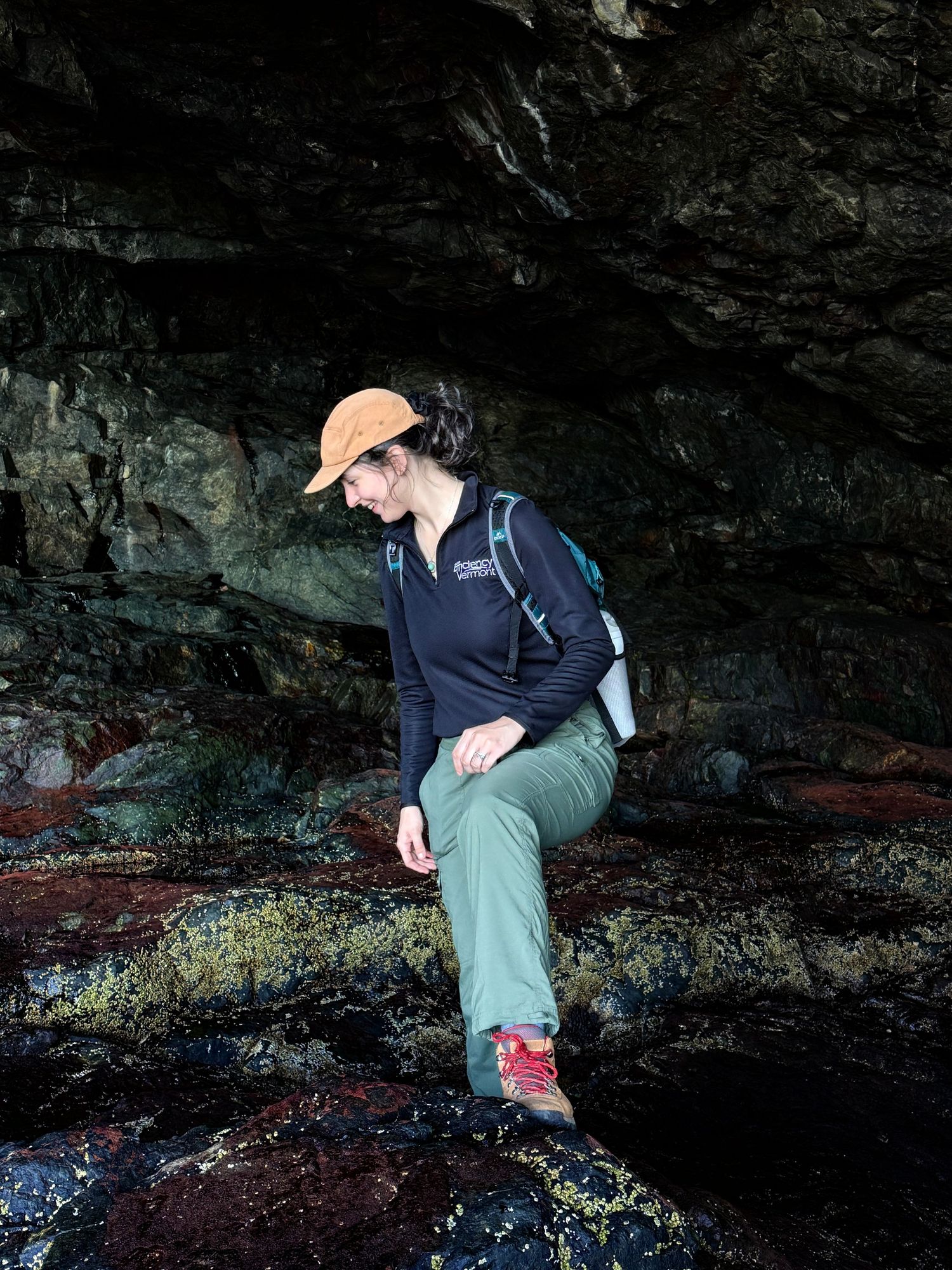 Woman in orange cap sitting on mossy rocks inside a dark cave during outdoor hiking adventure.