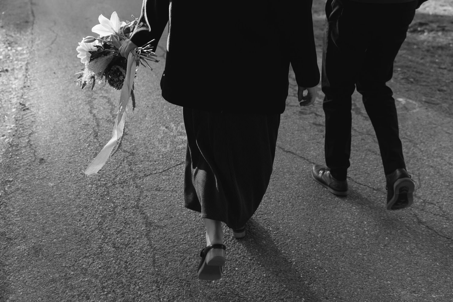Black and white photo of couple walking on road, woman in long skirt carrying a bouquet of sunflowers.