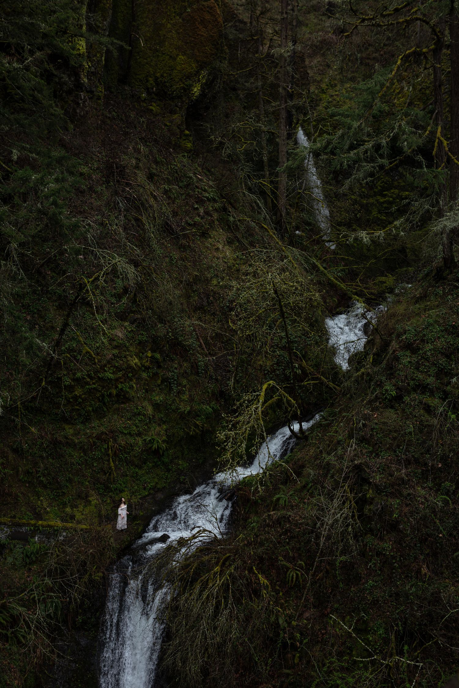 Woman in white dress standing beside rushing waterfall surrounded by dense mossy green forest.