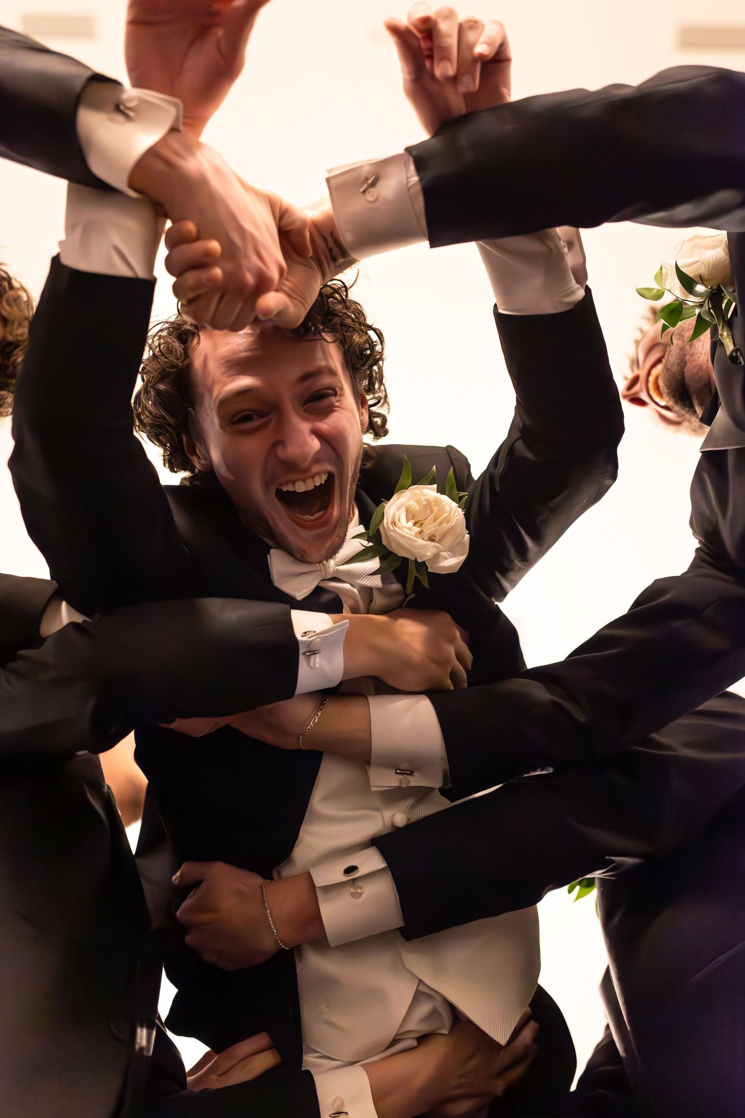 A person in a tuxedo celebrates joyfully while being lifted by others at a formal event.
