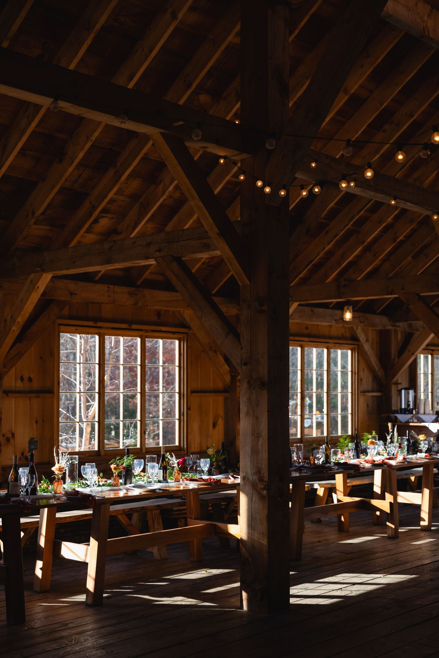 A rustic barn venue with wooden beams and string lights illuminating elegant table settings by large windows.