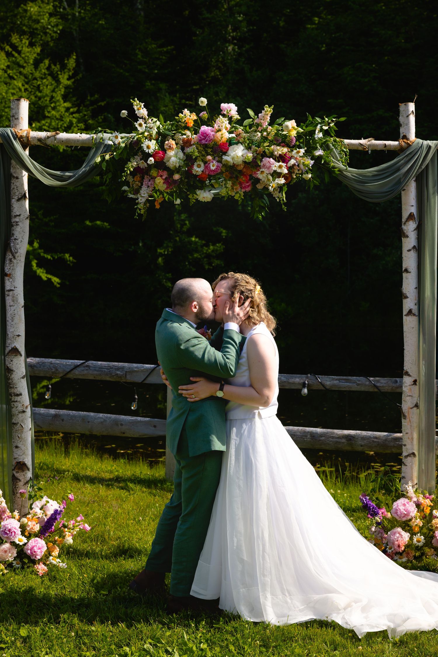 A romantic outdoor wedding ceremony takes place under a floral-adorned arbor in a sunlit meadow surrounded by trees.