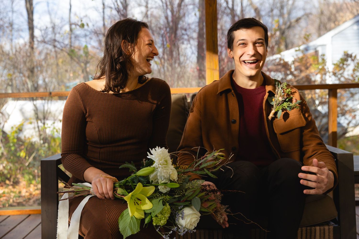 Two people sit on a porch together laughing and enjoying a sunny autumn day while holding white flowers.