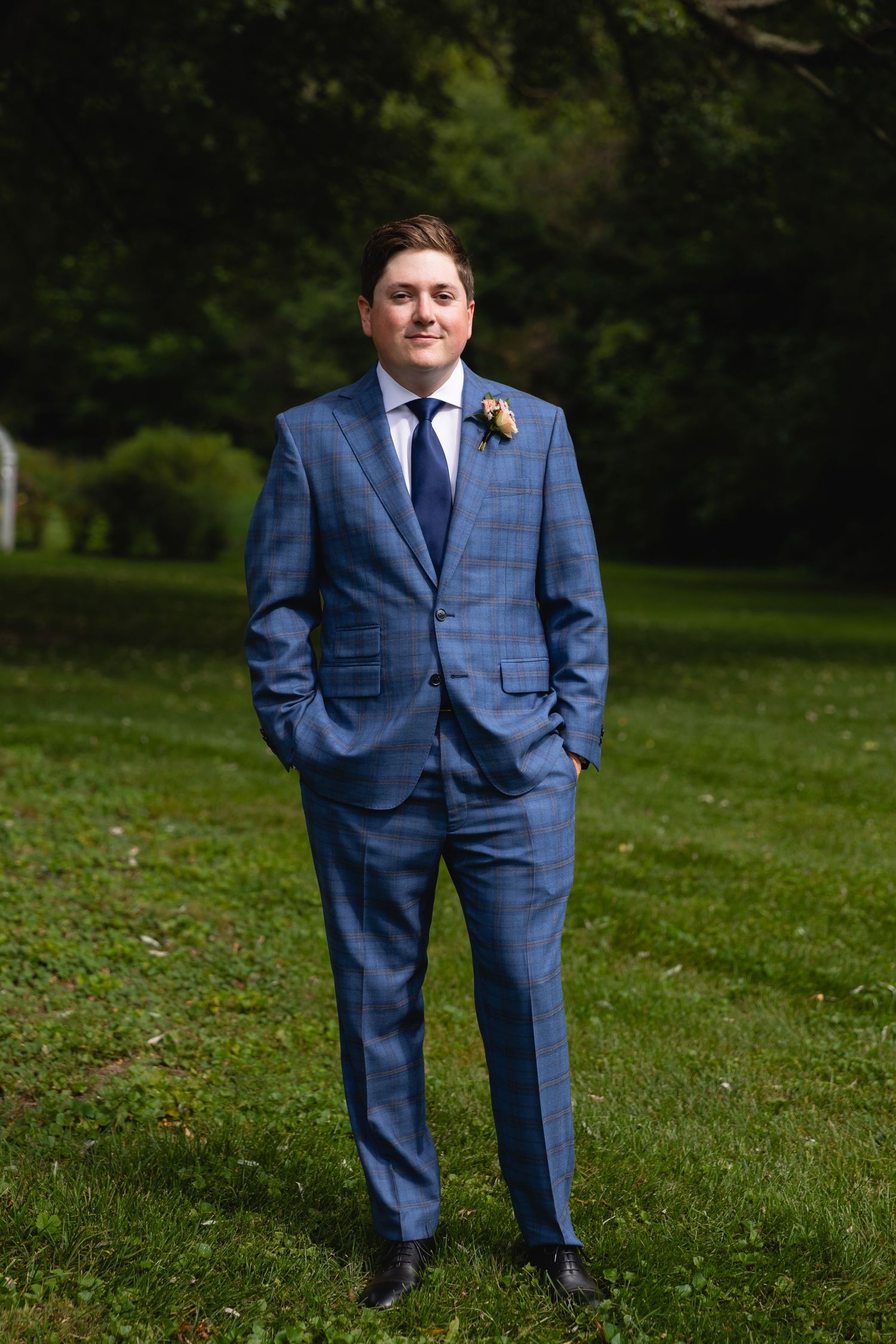 Groom poses outdoors in tailored blue windowpane suit with hands in pockets and boutonniere.