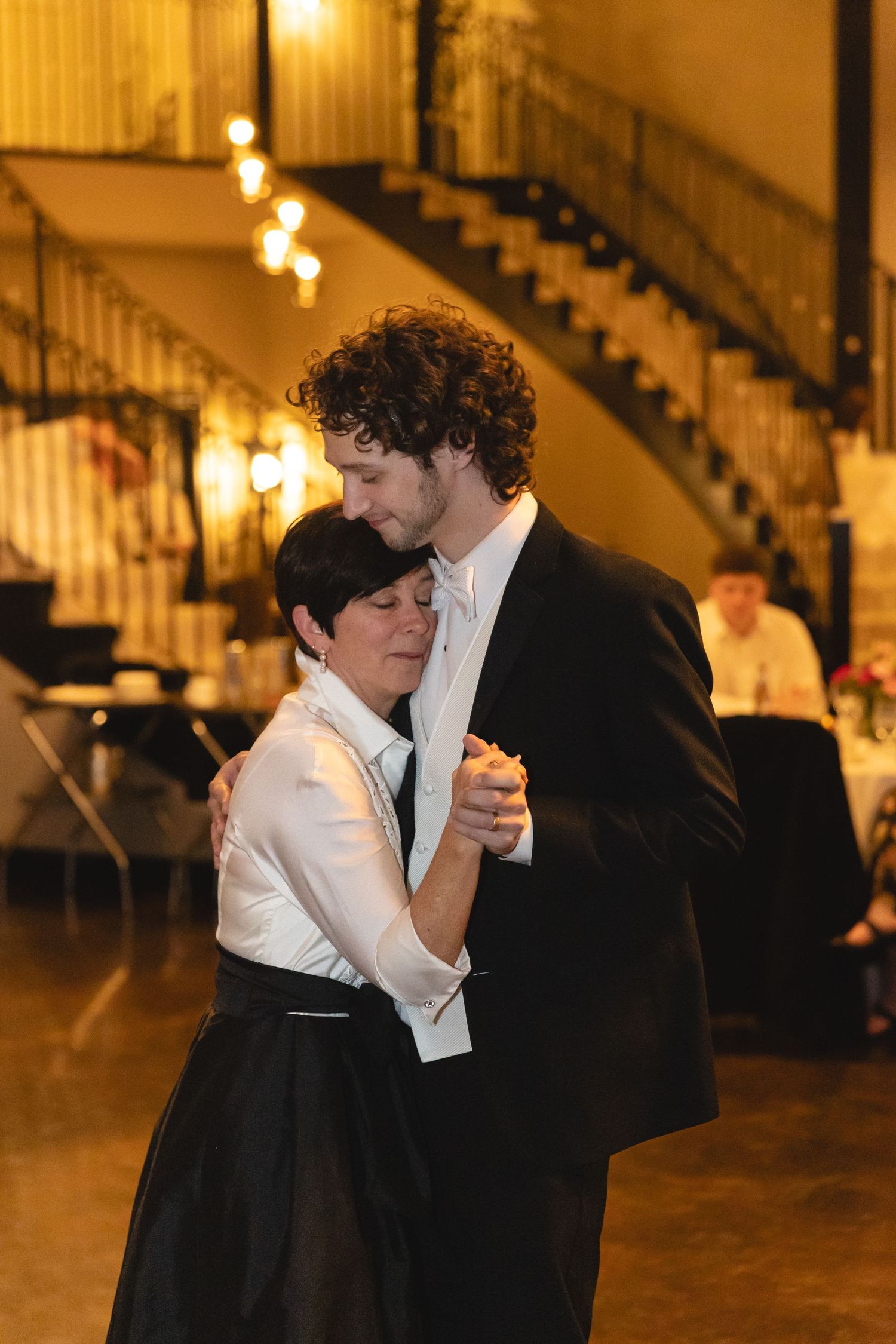 Couple shares intimate slow dance at elegant venue with curved staircase and warm lighting in background.