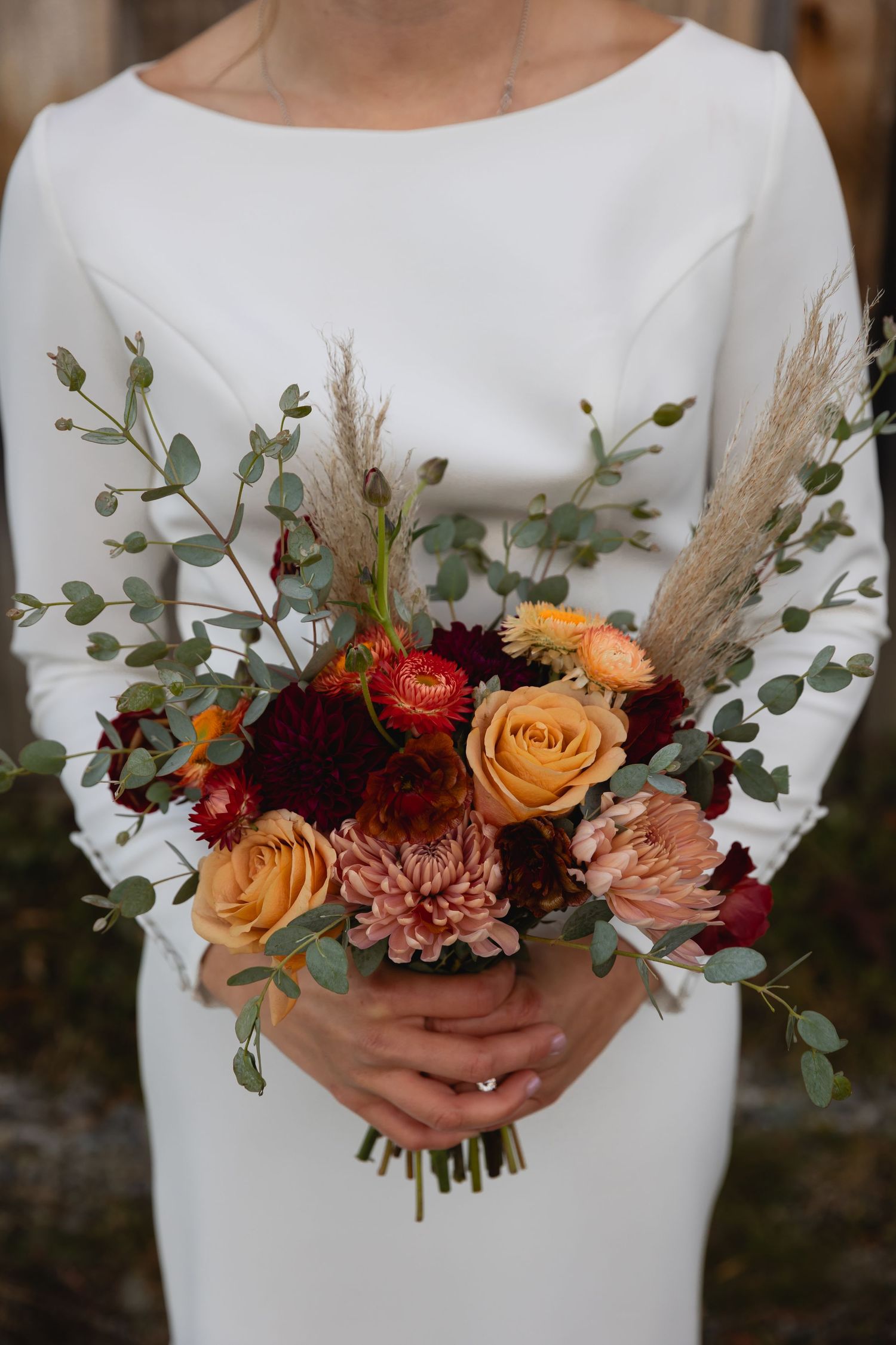 A stunning fall wedding bouquet with peach roses, burgundy dahlias, dried pampas grass and eucalyptus against a white dress.
