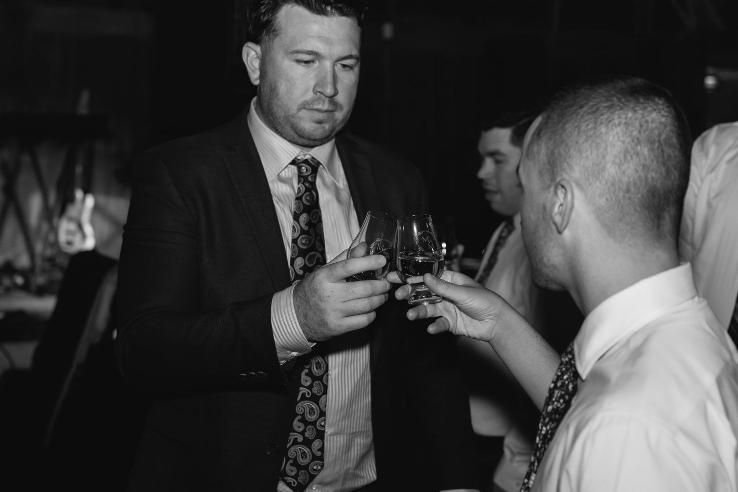 Two men in formal attire toast with glasses in a dimly lit venue, captured in black and white photography.