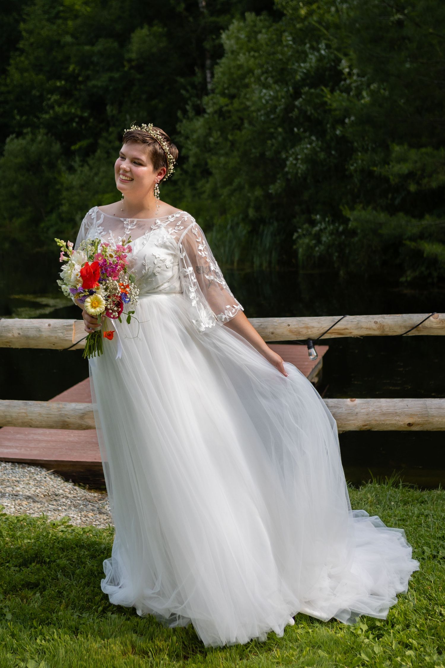Bride in ethereal white gown holds vibrant wildflower bouquet against green forest backdrop.