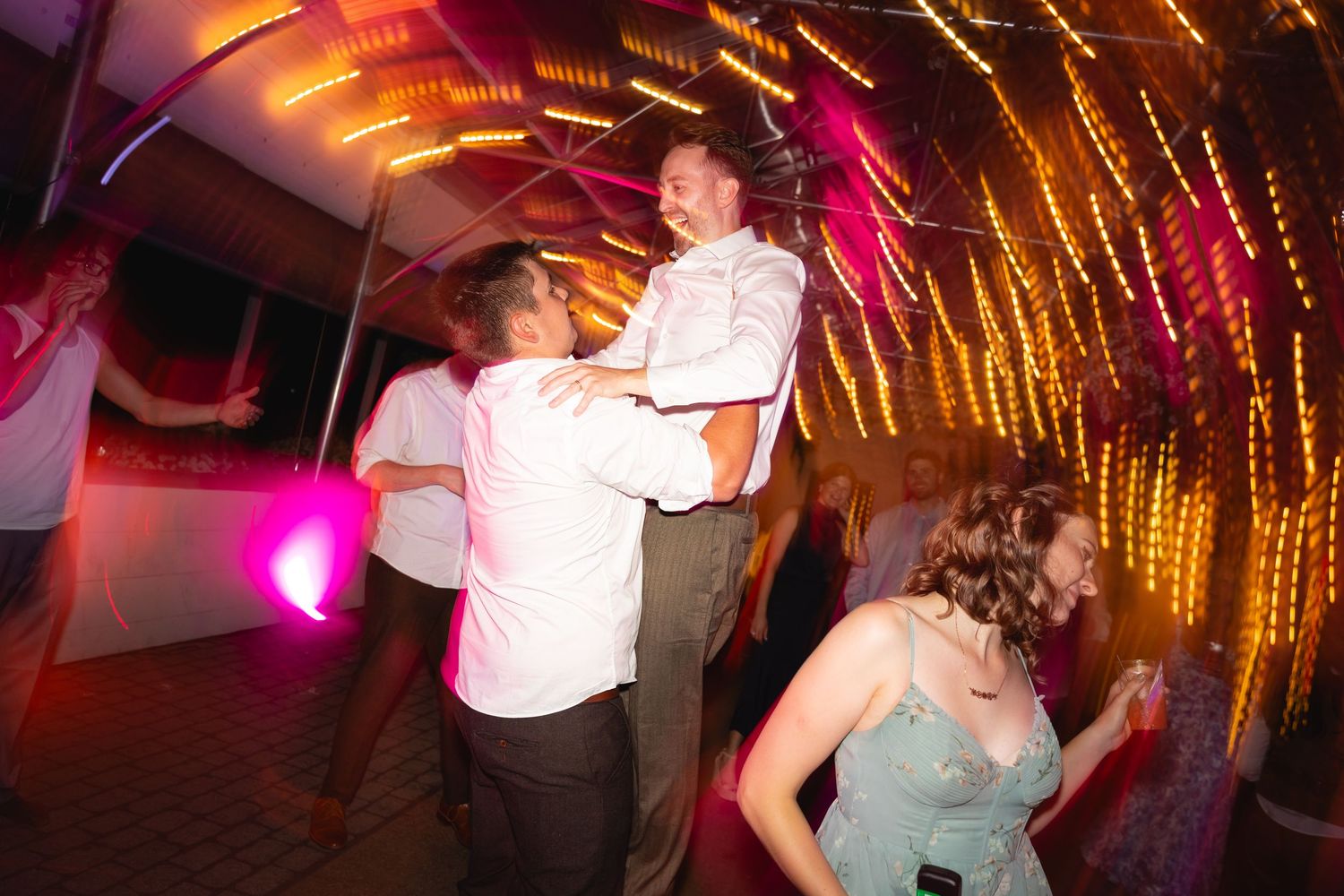 Wedding guests dancing under glowing string lights with pink and amber lighting creating a romantic ambiance.