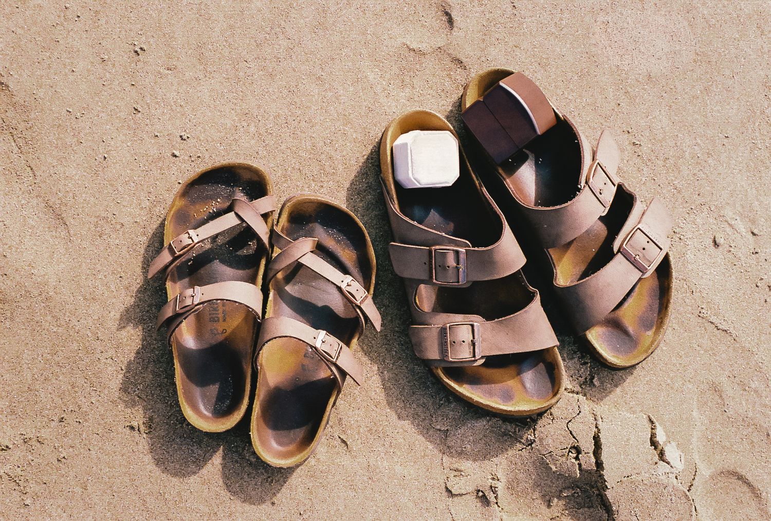 Two pairs of brown leather sandals with buckle straps sitting on sandy beach in natural sunlight.