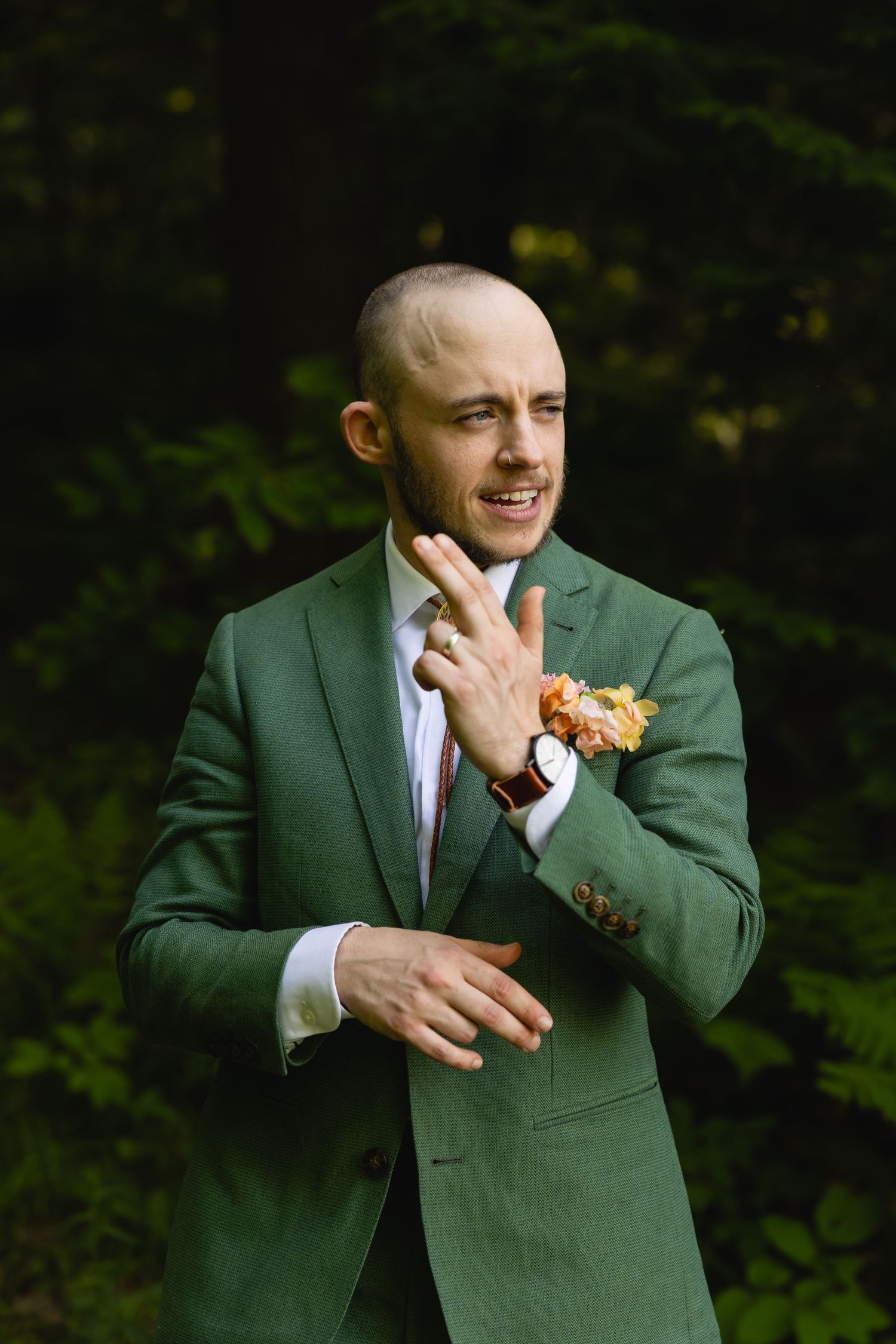 A groom in a forest green suit jacket with a floral boutonniere gestures expressively against a dark natural background.
