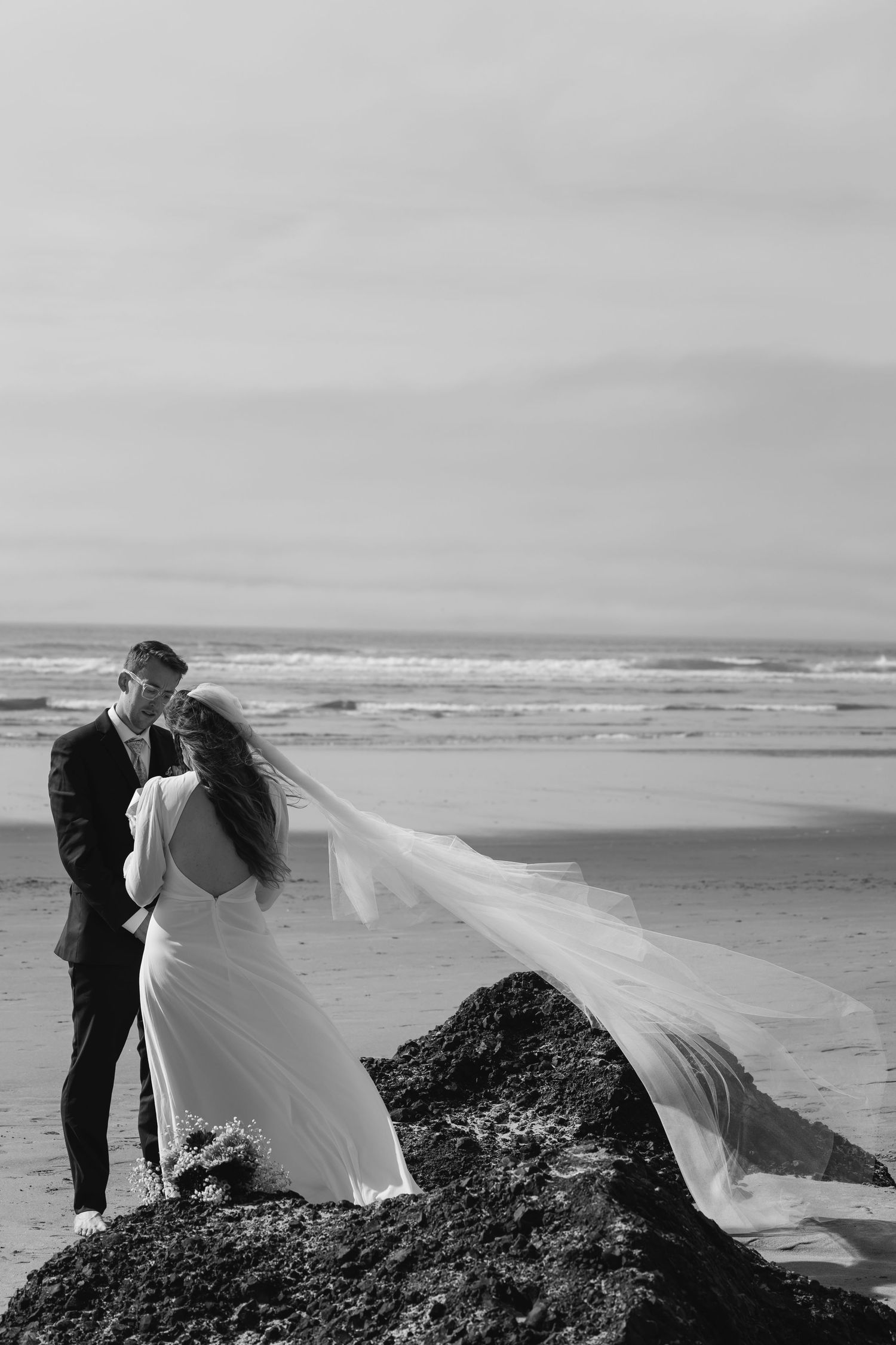 A romantic black and white wedding photoshoot on a dramatic coastal cliff with a flowing veil catching the ocean breeze.