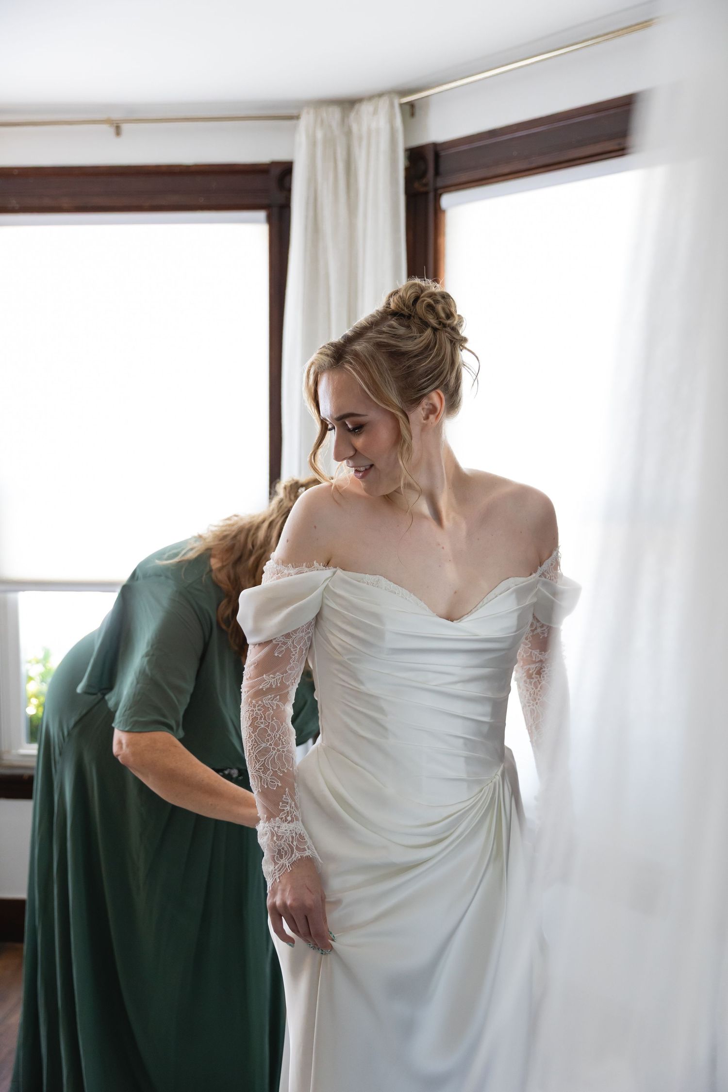 A bride in a white wedding dress stands near a window while someone in emerald green helps with final preparations.