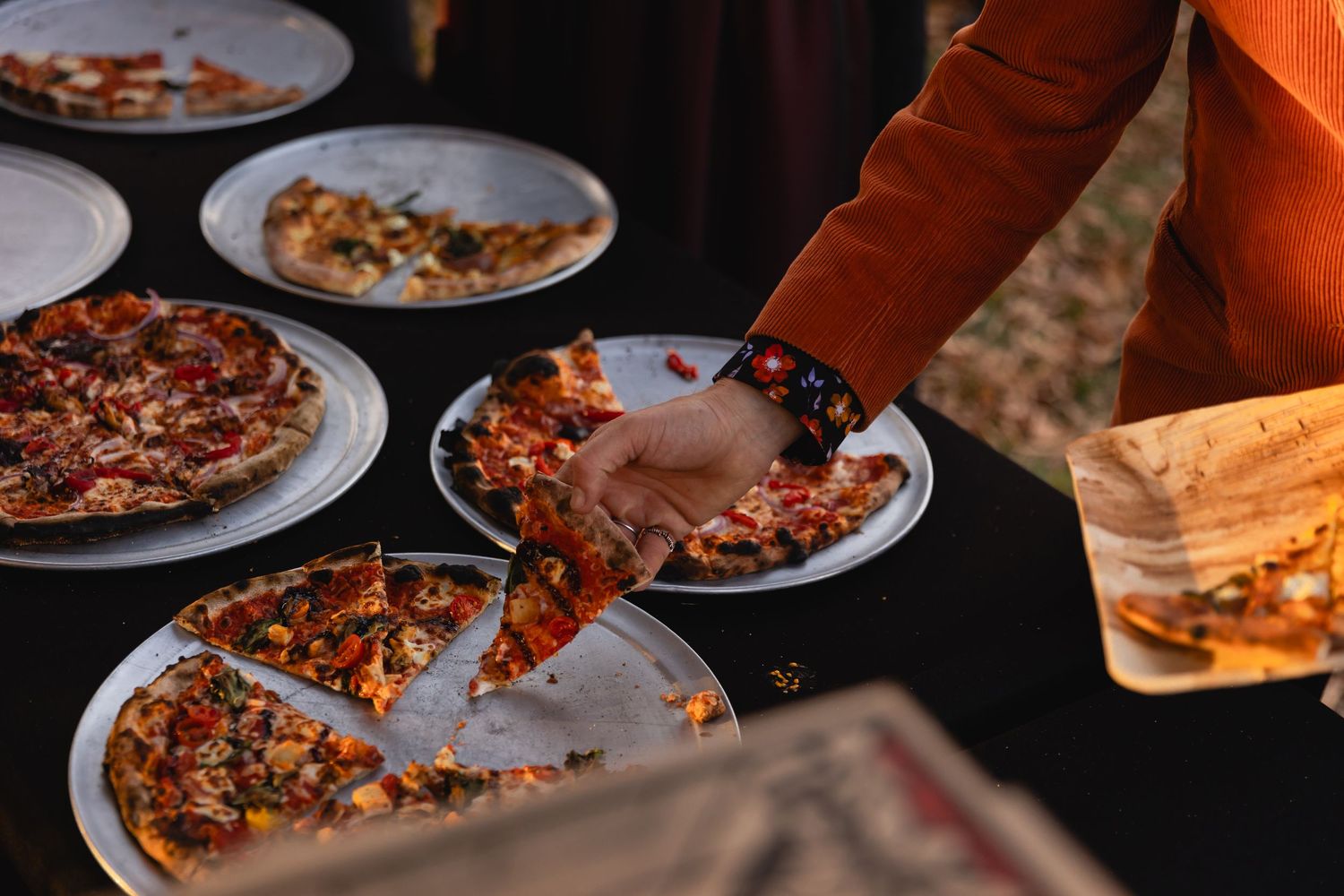 Hands serving slices of pizza on plates during a casual outdoor dining gathering.
