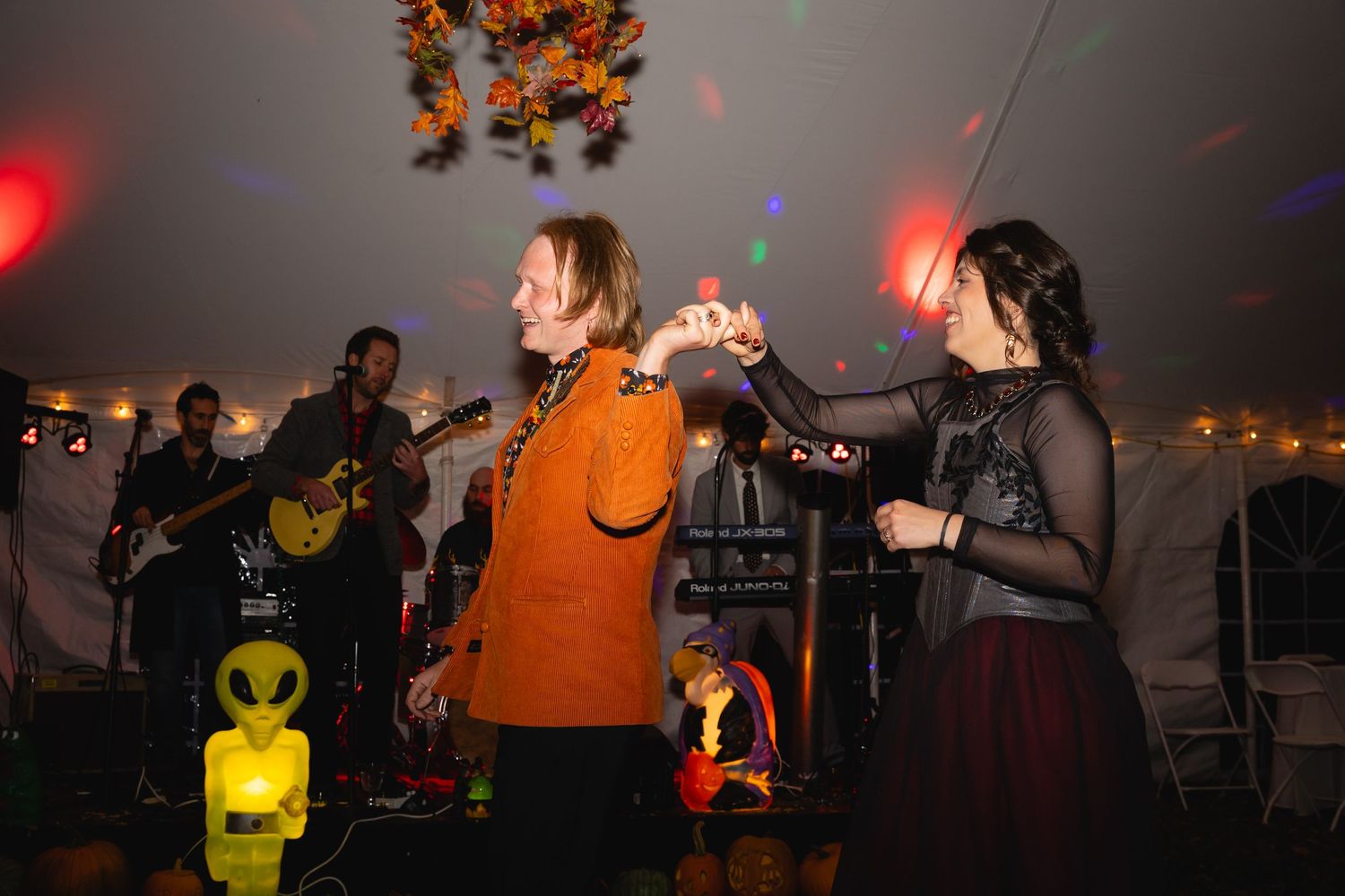 People dance and celebrate at a dimly lit party with colorful lights and an inflatable alien decoration visible.