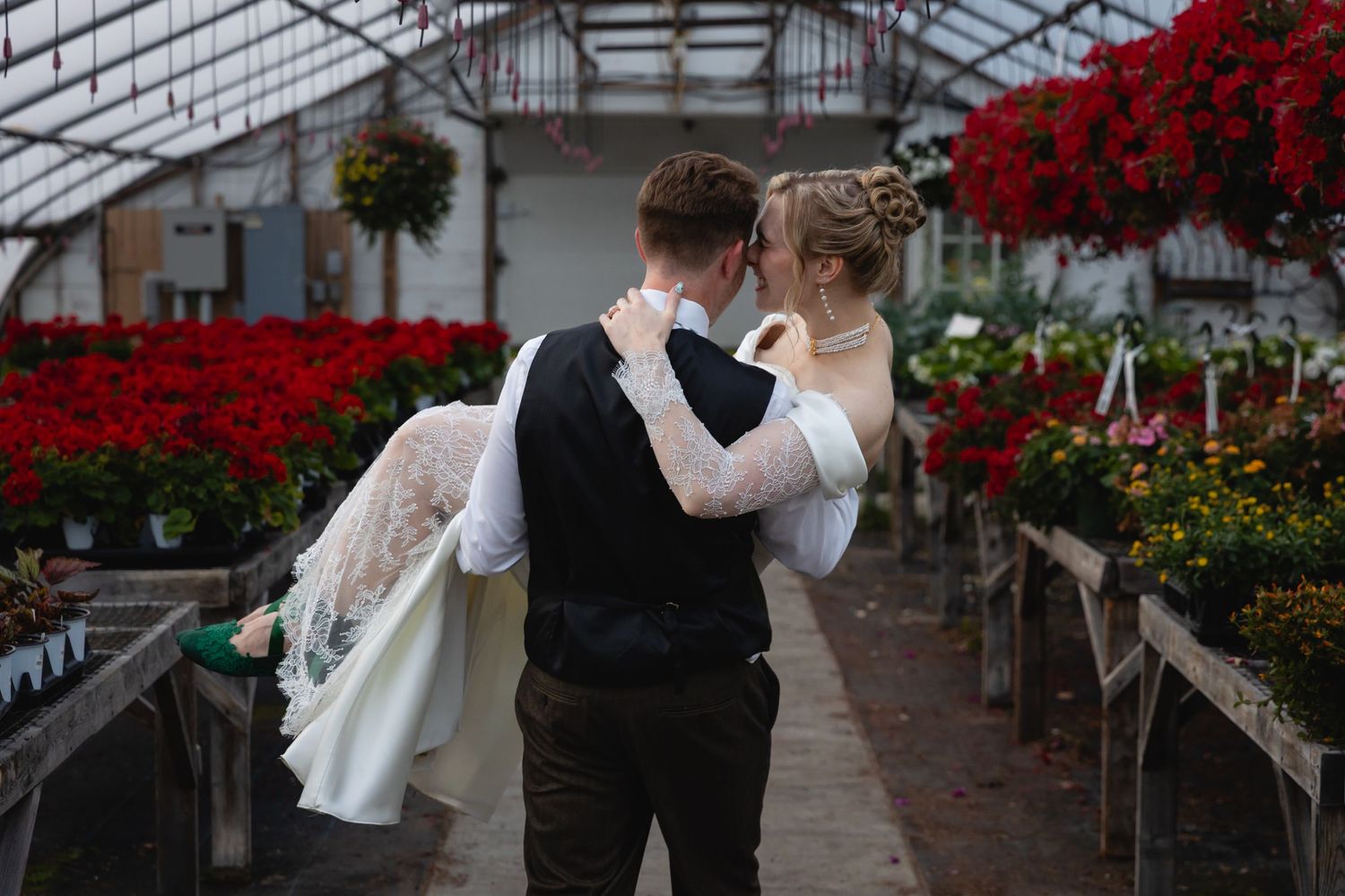 Couple embraces in a greenhouse surrounded by vibrant red flowers and hanging plants.