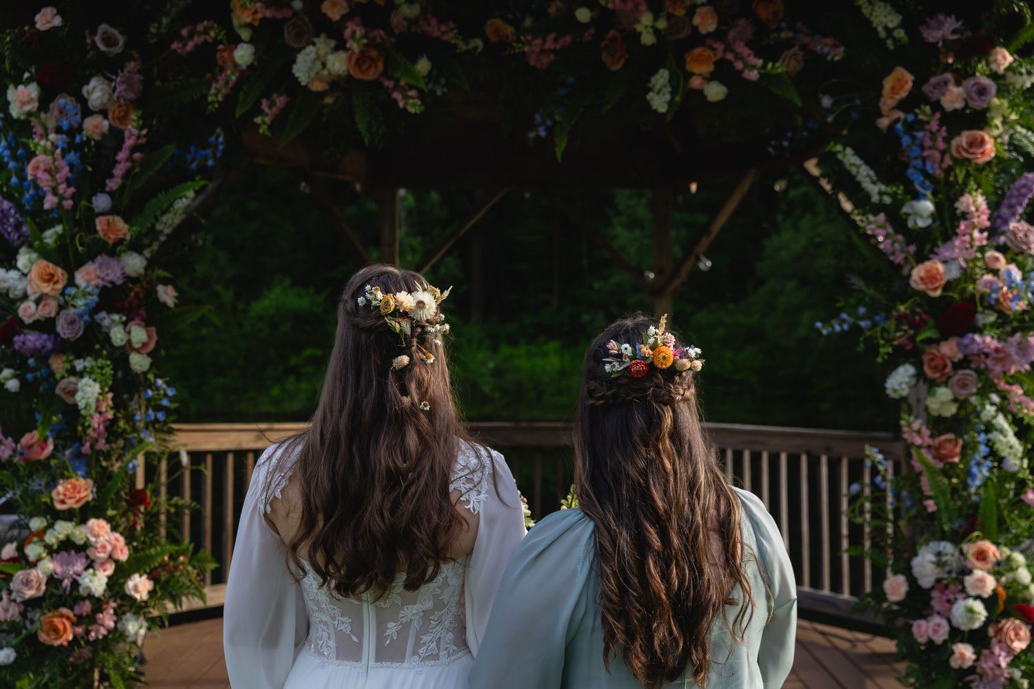 Two brides standing before colorful floral arch with flower crowns in their hair.