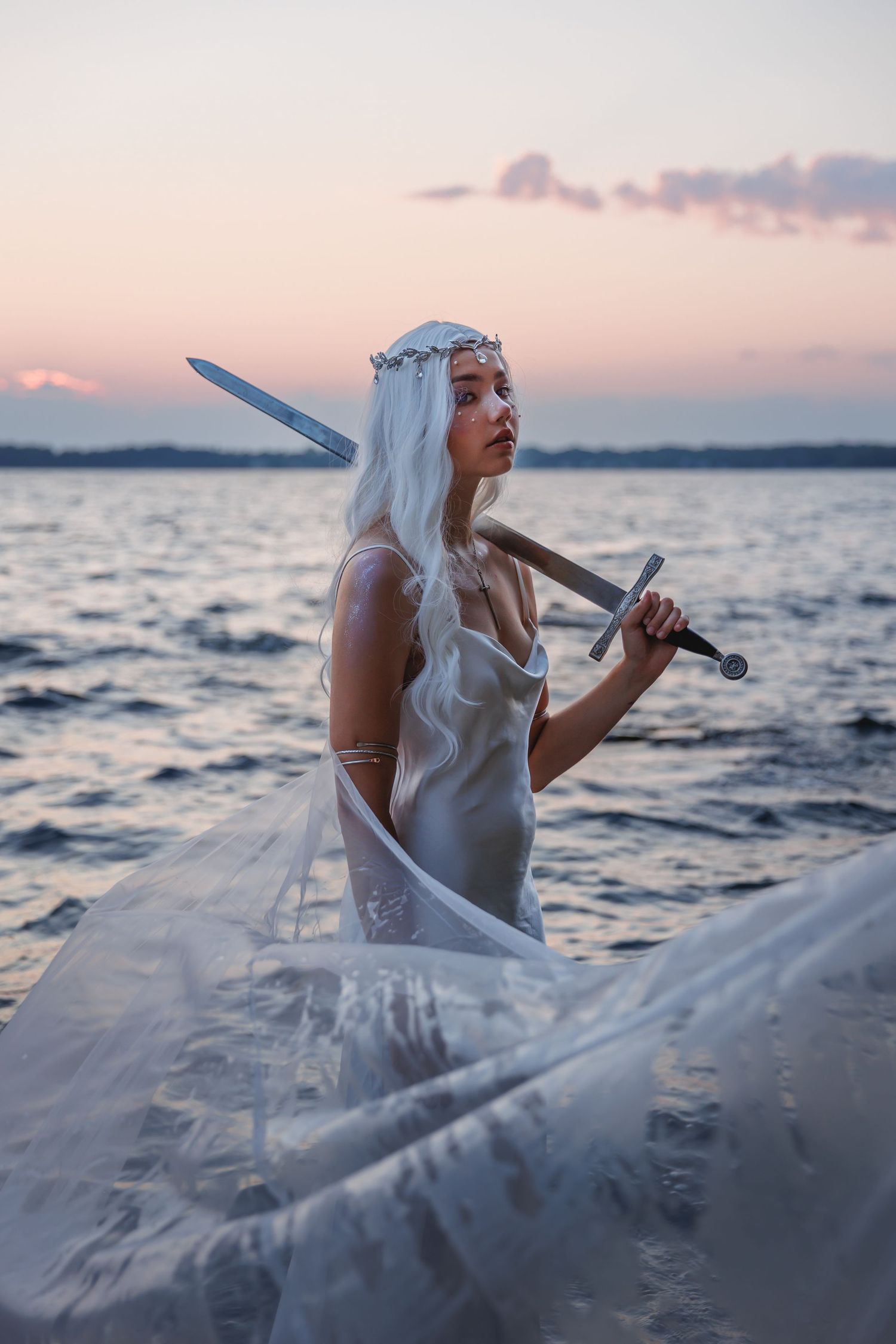 White-haired woman in flowing white gown holds sword while standing in lake at sunset.