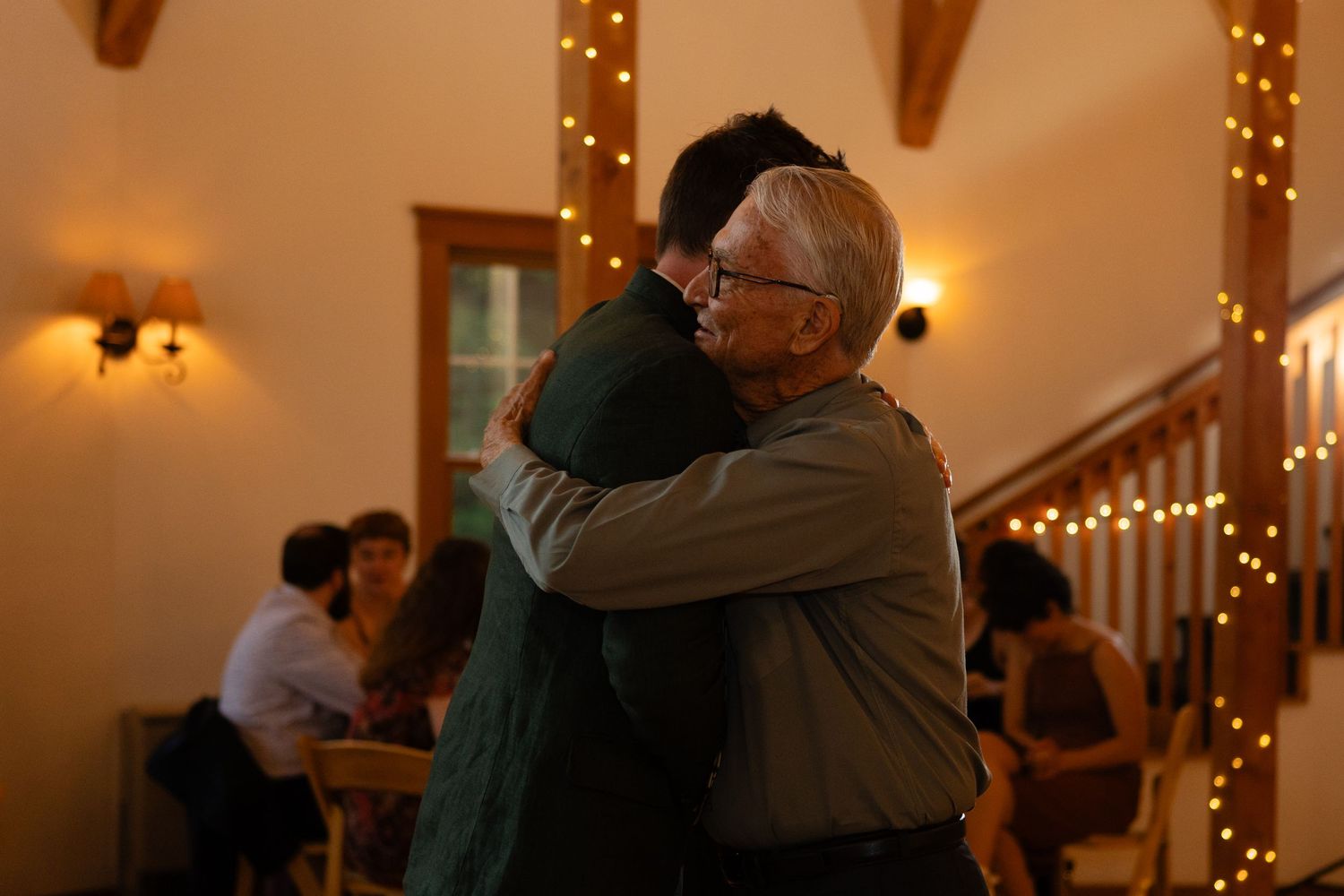 Two people share an emotional embrace during a reception with twinkling lights in the background.