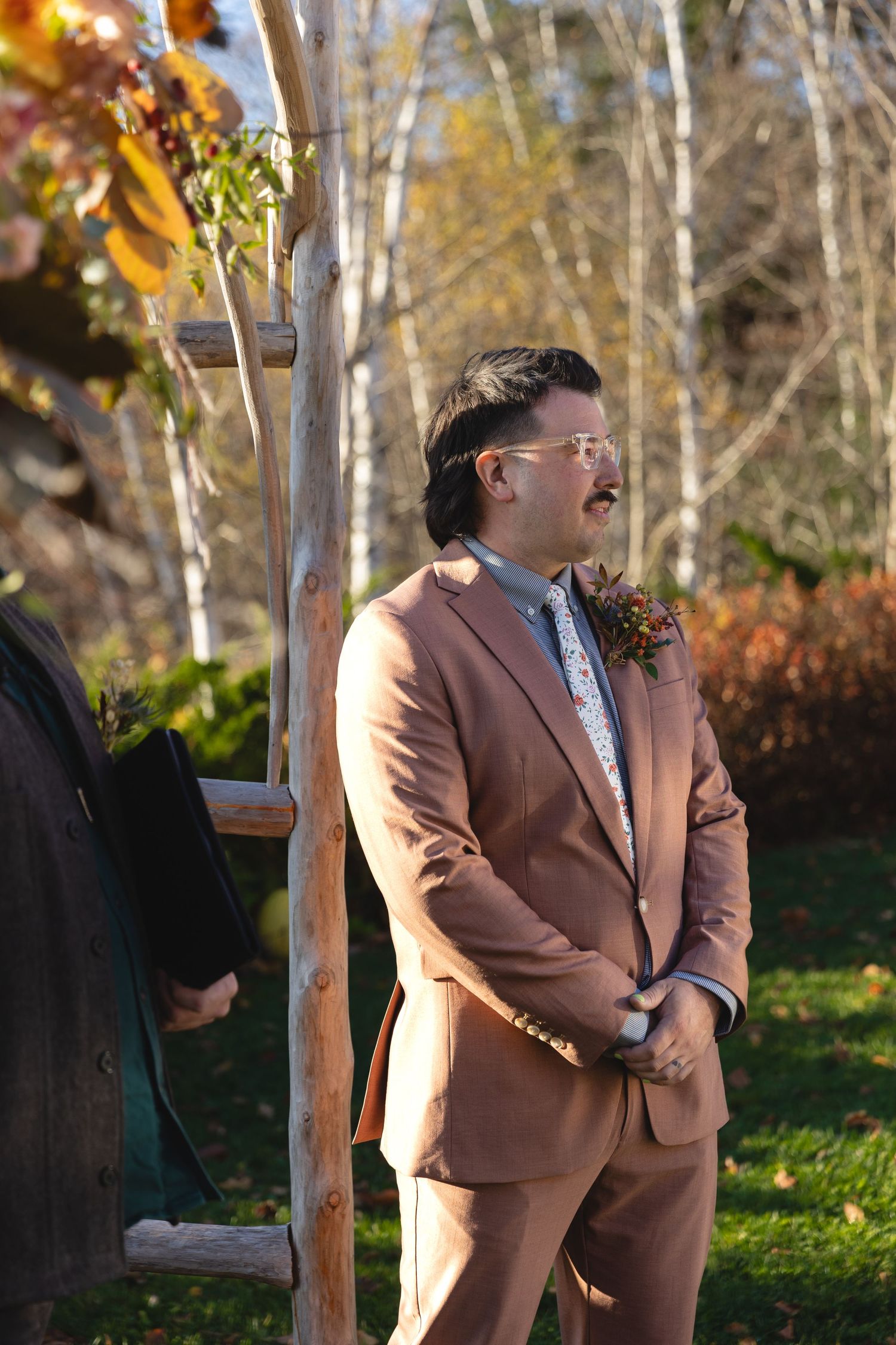 Wedding participant in light brown suit standing outdoors during fall ceremony.