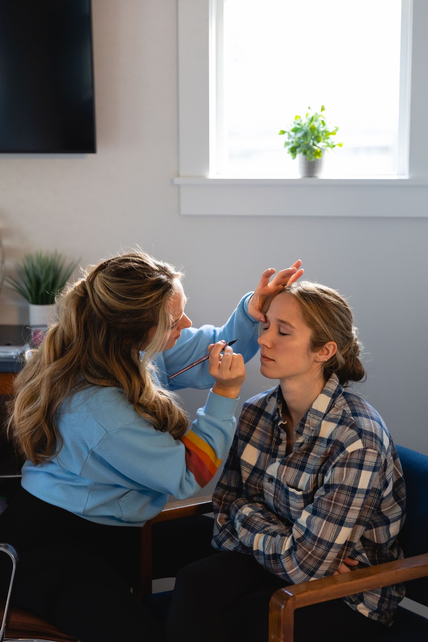 Professional makeup artist applying eye makeup to client in home studio with natural window lighting and potted plants.