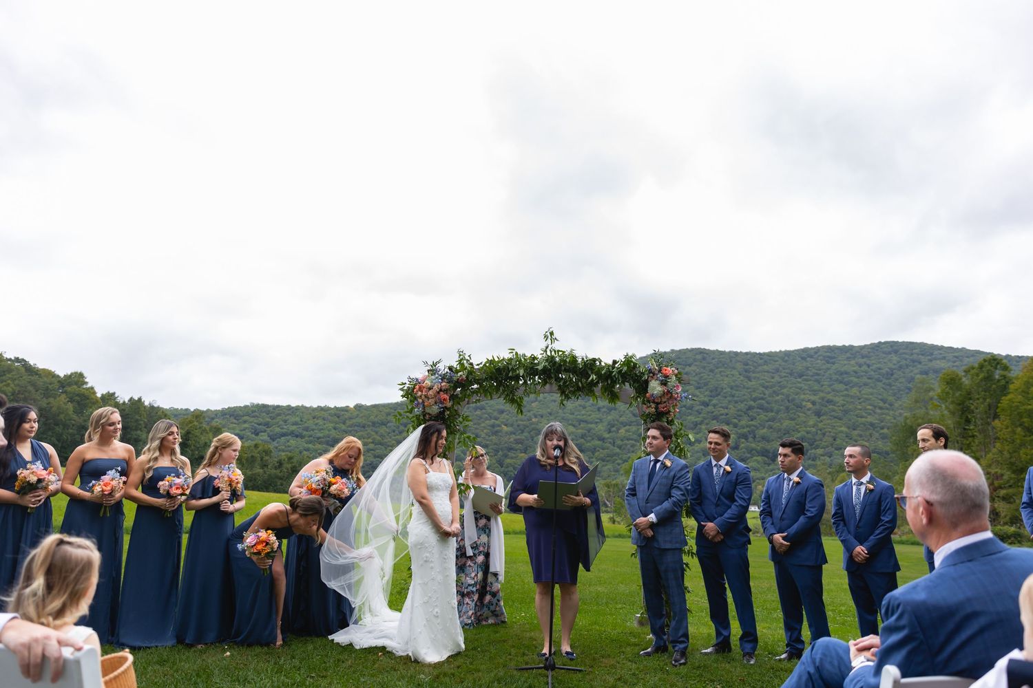 A wedding ceremony takes place outdoors on a hilltop with guests in navy blue and bridesmaids in dark blue dresses.
