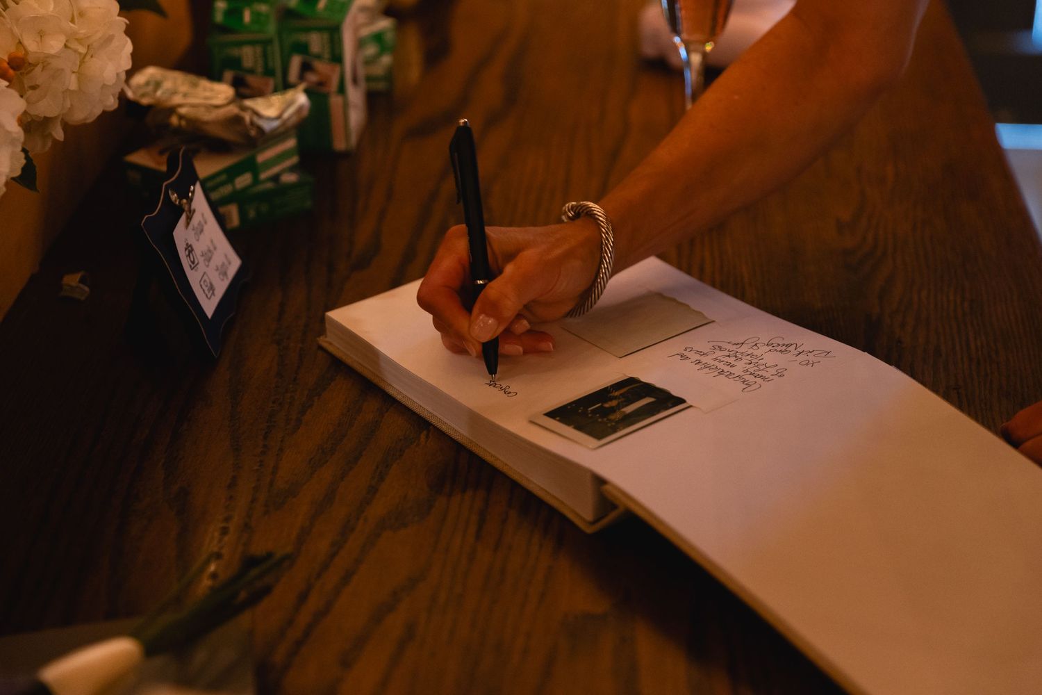 Hand holding pen writing in a book on dark wooden table in warm lighting.